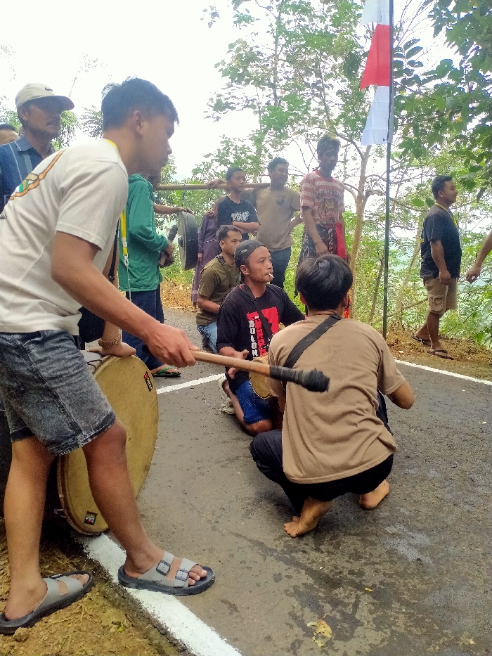 This image depicts an outdoor scene featuring approximately ten adult males on an asphalt road in Ngargoyoso, Indonesia. In the left foreground, a male wearing a white t-shirt and grey denim shorts stands beside a large, light-colored frame drum, holding two drumsticks in his right hand. To his immediate right, a male in a black t-shirt and black baseball cap squats, holding a drumstick over a smaller frame drum, with a lit cigarette in his mouth. Behind him and slightly to his right, another male squats, viewed from the back, wearing a brown t-shirt and dark pants, also holding a drumstick. Among the standing individuals in the mid-ground, one male carries a large, dark gong suspended from a bamboo pole across his right shoulder. Another male with disheveled hair, wearing a striped t-shirt and red sarong, stands further right. An Indonesian red and white flag is affixed to a pole on the far right of the road. The asphalt road features a distinct white painted line along its left edge. The background is dominated by dense green foliage and trees, indicating a natural or elevated rural environment.
