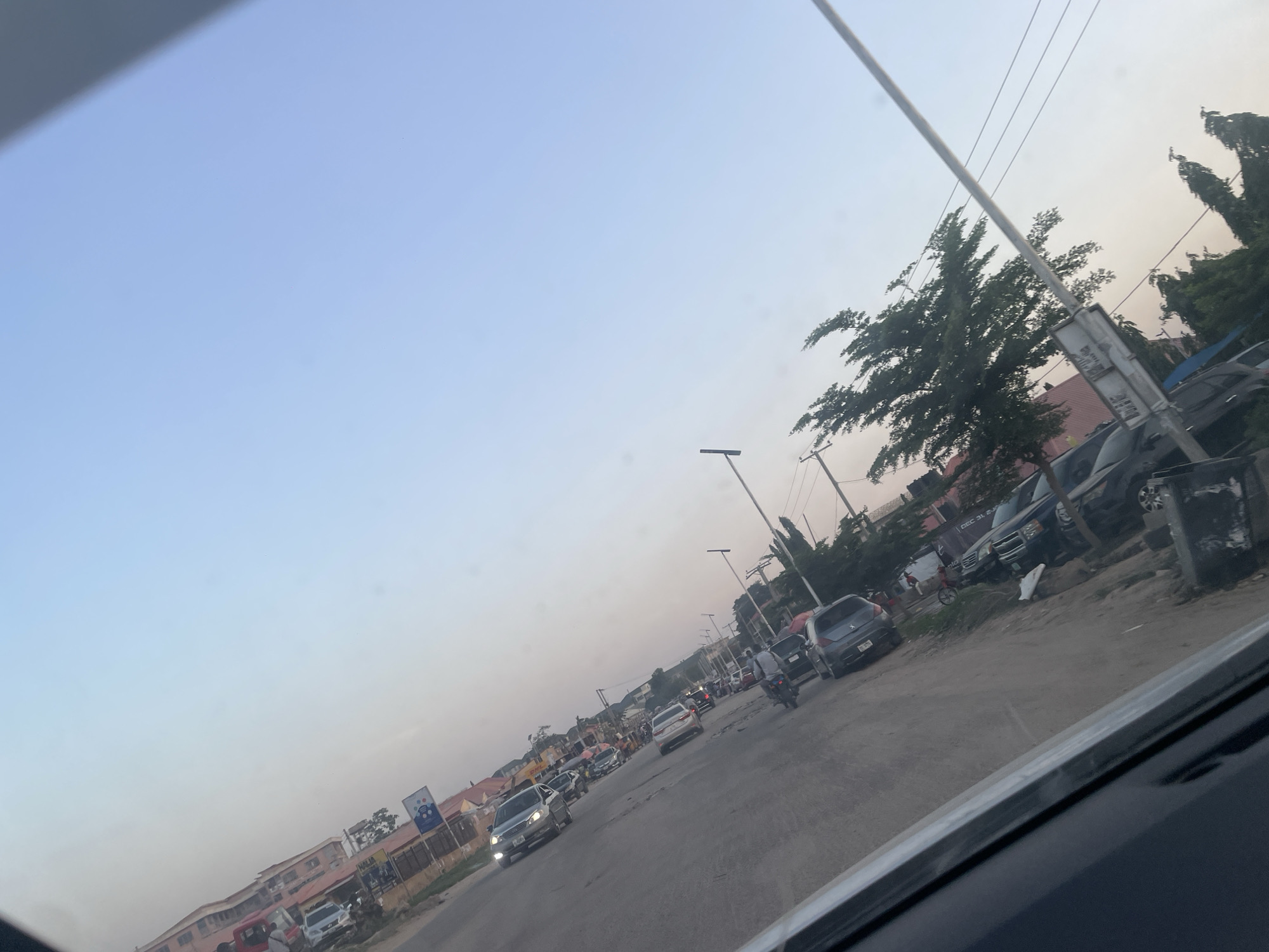The image depicts a street scene in Abuja, Nigeria, likely captured from inside a vehicle. The road is paved and appears to be moderately busy, with a mix of cars, motorcycles, and other vehicles traveling in both directions. Some vehicles are parked on the side of the road. Buildings and trees line the street, and utility poles with power lines are visible. The sky is light blue, suggesting either early morning or late afternoon. There are a few pedestrians and a bicyclist in the scene. The image has been taken from a low angle, possibly from inside a car.