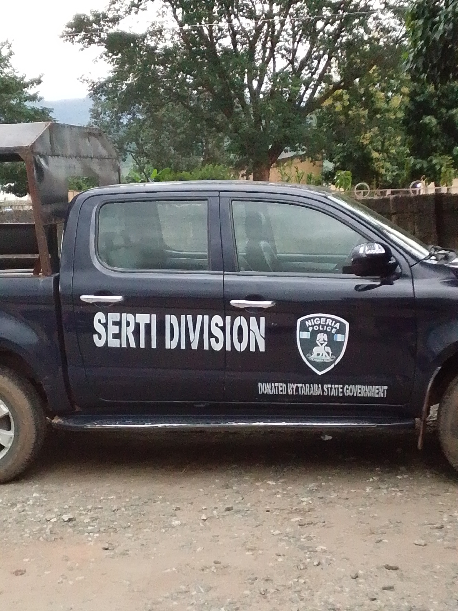 The image shows a dark blue pickup truck parked on a gravelly surface. The truck has "SERTI DIVISION" printed in large white letters on its side. To the right of the text is a shield-shaped emblem with "NIGERIA POLICE" written on it. Below the emblem and the "SERTI DIVISION" text, smaller white letters read "DONATED BY TARABA STATE GOVERNMENT". The truck has a metal cage-like structure in its bed. The environment appears to be outdoors with trees and greenery visible in the background. The overall scene suggests the truck is located in or associated with the Serti division in Taraba State, Nigeria.