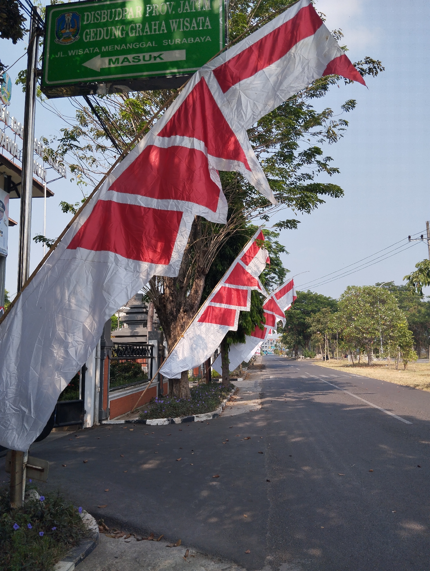 A street view shows a road leading into a residential or government complex in Surabaya, Indonesia. Indonesian flags, featuring red and white horizontal stripes, are hung on poles along the side of the road. The flags are large and appear to be made of fabric. A green sign with white lettering is visible on the left side of the image. The sign reads "DISBUDPAR PROV. JATIM GEDUNG GRAHA WISATA" and "JL. WISATA MENANGGAL SURABAYA MASUK" with an arrow pointing left. The road is paved and has a white line marking the center. Trees and vegetation line the sides of the road. The sky is clear and blue. There are no people clearly visible in the image. The image appears to have been taken during the daytime.
