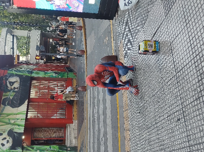 Spiderman kneels on a tiled sidewalk next to a yellow and blue box. A yellow line separates the sidewalk from the street, where a mural of pandas adorns a red building. People walk in the background. A digital screen looms overhead.