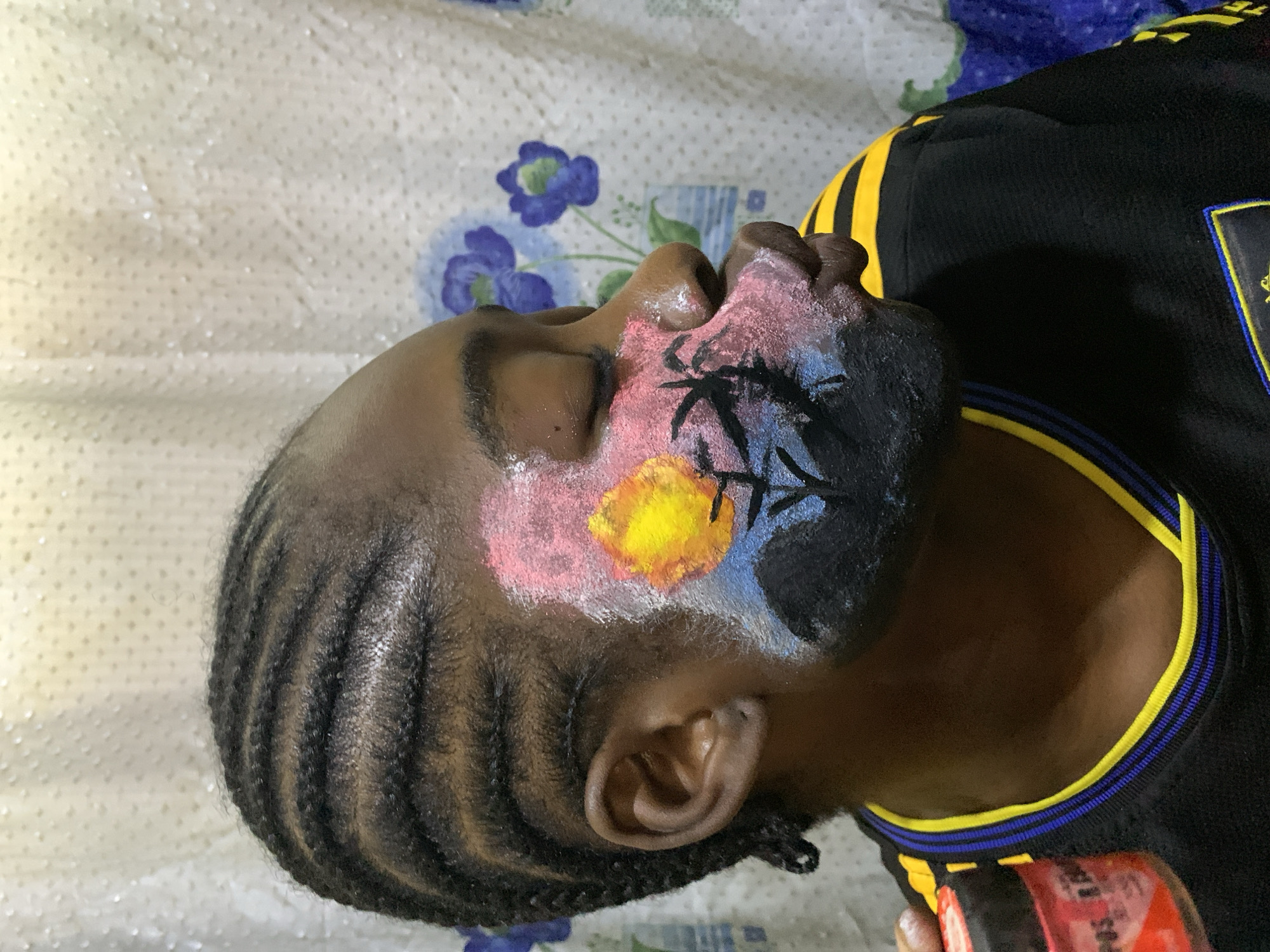The image shows a close-up, sideways view of a person's head, with their face painted. The person has dark skin and their hair is styled in braids. The face paint includes a yellow and orange sun or celestial body, with pink and blue colors surrounding it. There are also black lines painted on the face, resembling branches or abstract shapes. The person is wearing a black shirt with yellow and blue stripes around the collar.
The background of the image is a light-colored cloth with a subtle pattern of raised dots and some blue floral designs. In the bottom right corner, a hand is visible holding a red container with some text on it, though the text is not clearly legible.
The lighting suggests it might be daytime, but no specific time or weather conditions can be determined. The setting appears to be indoors, given the fabric background. The expression on the person's face is neutral, with their eyes closed. The overall impression is that of face painting being applied or recently completed, possibly for an event or as a form of artistic expression. The location context provided is Rafin Par, Nigeria.