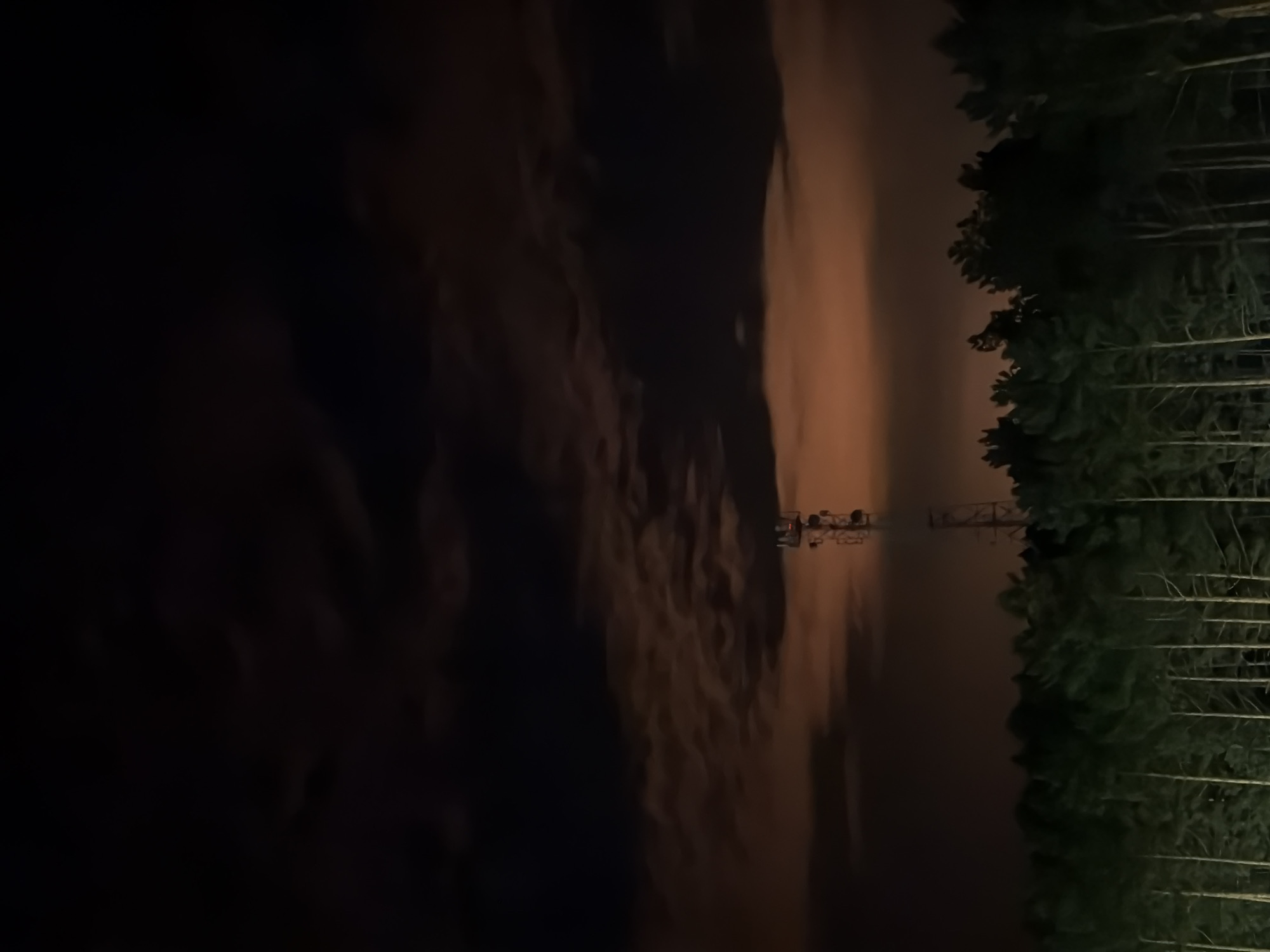The image depicts a nighttime scene with a cell tower silhouetted against a hazy, orange-tinted sky, possibly illuminated by distant city lights or atmospheric conditions. Below the sky is a dense line of dark green pine trees, appearing to be a forest. The cell tower is tall and metallic, with visible antennas at its peak. The foreground consists of the tops of the pine trees. The overall impression is one of a remote or semi-rural area at night. The location is specified as the city of Tsiolkovsky, Russia, which is known for its spaceport and associated infrastructure.