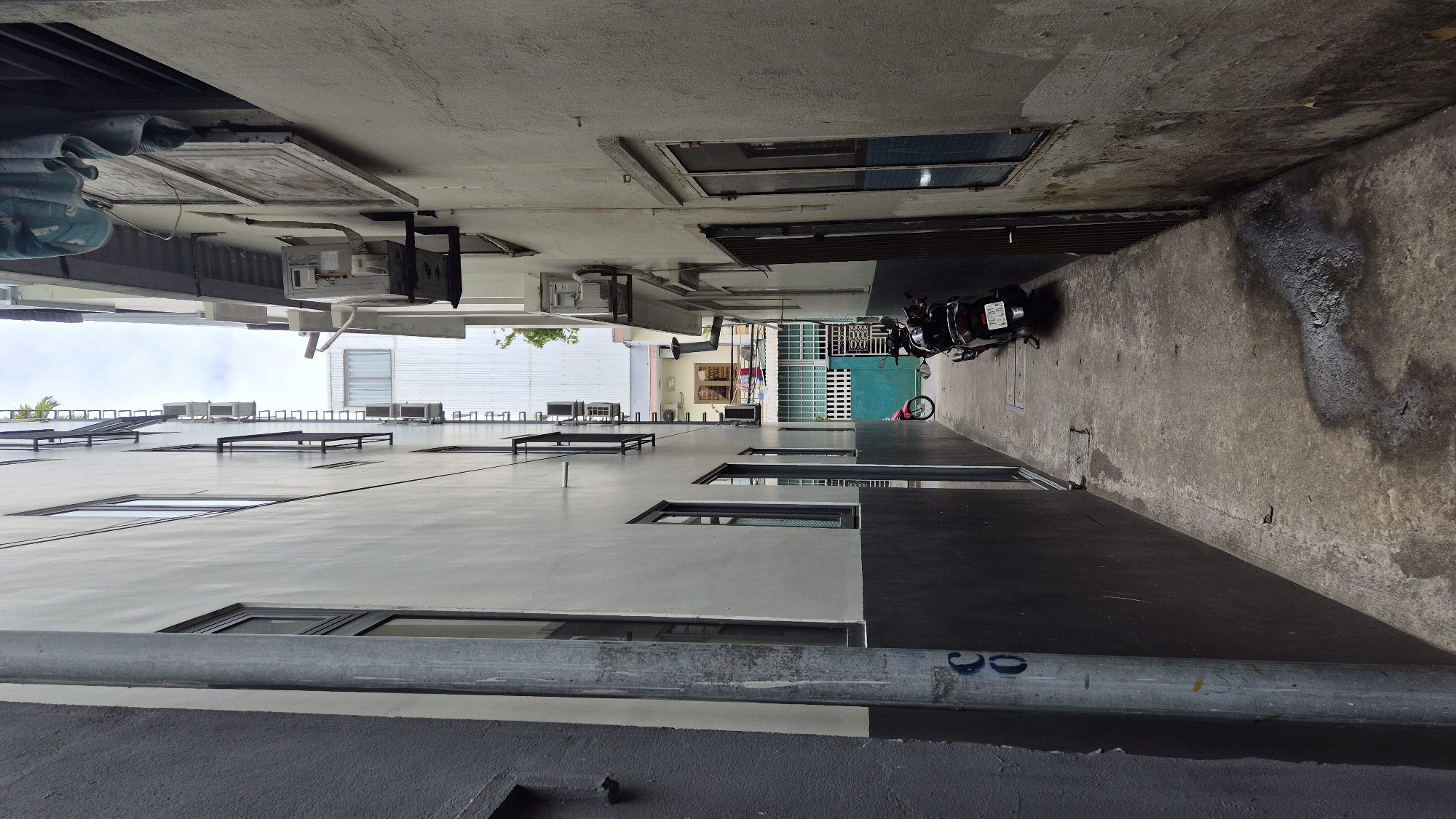 The image depicts a narrow alleyway between two buildings in Đà Nẵng, Vietnam. The alley floor is paved with concrete, showing signs of wear and water stains. On the left, a grey building with a dark grey lower section runs along the alley. A metal pipe with some markings, including the letter "C" and the number "0", is visible on this building's wall. The building on the right is lighter in color and appears to be older, with visible water damage and some grime on its exterior walls.
Several objects are present in the alley. Two motorcycles are parked towards the end of the alley. One is a black scooter, facing away from the camera, with a license plate that reads "74-D1 453.08". A red motorcycle is parked further down the alley, to the left of the black scooter. In the background, a teal-colored gate is visible, partially obscured by the motorcycles. Above the alley, the buildings rise, revealing multiple windows, air conditioning units, and some laundry hanging out to dry. The sky is overcast, suggesting a cloudy day. The overall impression is one of a functional, utilitarian space within a densely populated urban environment.