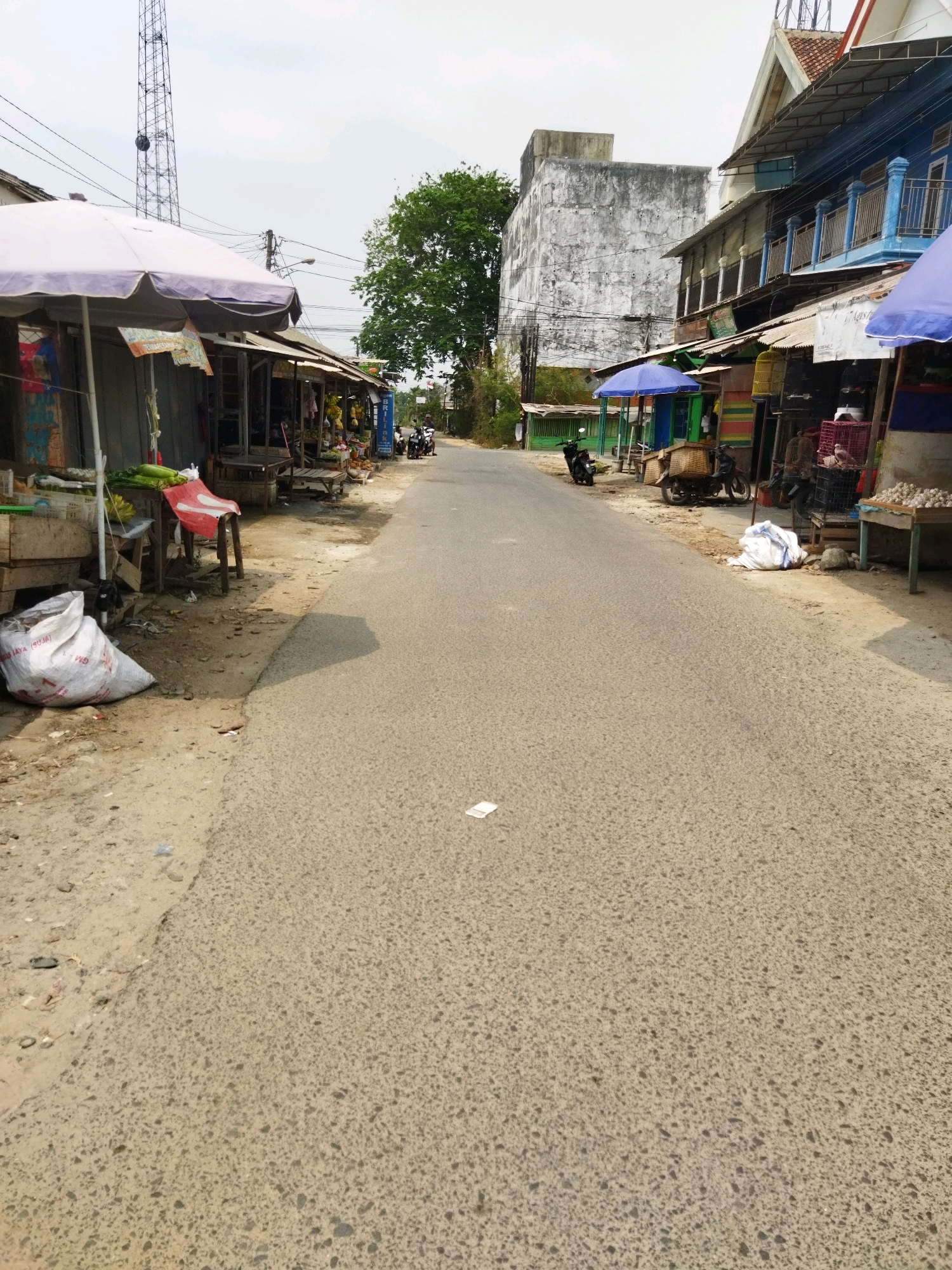 This is a medium-angle, full-shot, eye-level, outdoor, daytime photograph of a street scene in Banjar Agung, Indonesia. The street is paved with asphalt and is bordered by small market stalls on both sides. The stalls appear to be made of wood and corrugated metal, with some covered by umbrellas or awnings. On the left side of the street, there is a large white sack of what appears to be produce, possibly rice or potatoes, near a stall with green vegetables. A red chair is also visible at this stall. Further down the street on the left, a tall radio tower is visible. On the right side of the street, there are more market stalls selling various goods, including what appear to be birds in cages, vegetables, and other food items. Several motorcycles are parked along the side of the road, and a few people are seen in the distance, near the stalls. A tall, weathered concrete building stands behind the stalls on the right. The sky is overcast and the overall lighting is subdued.