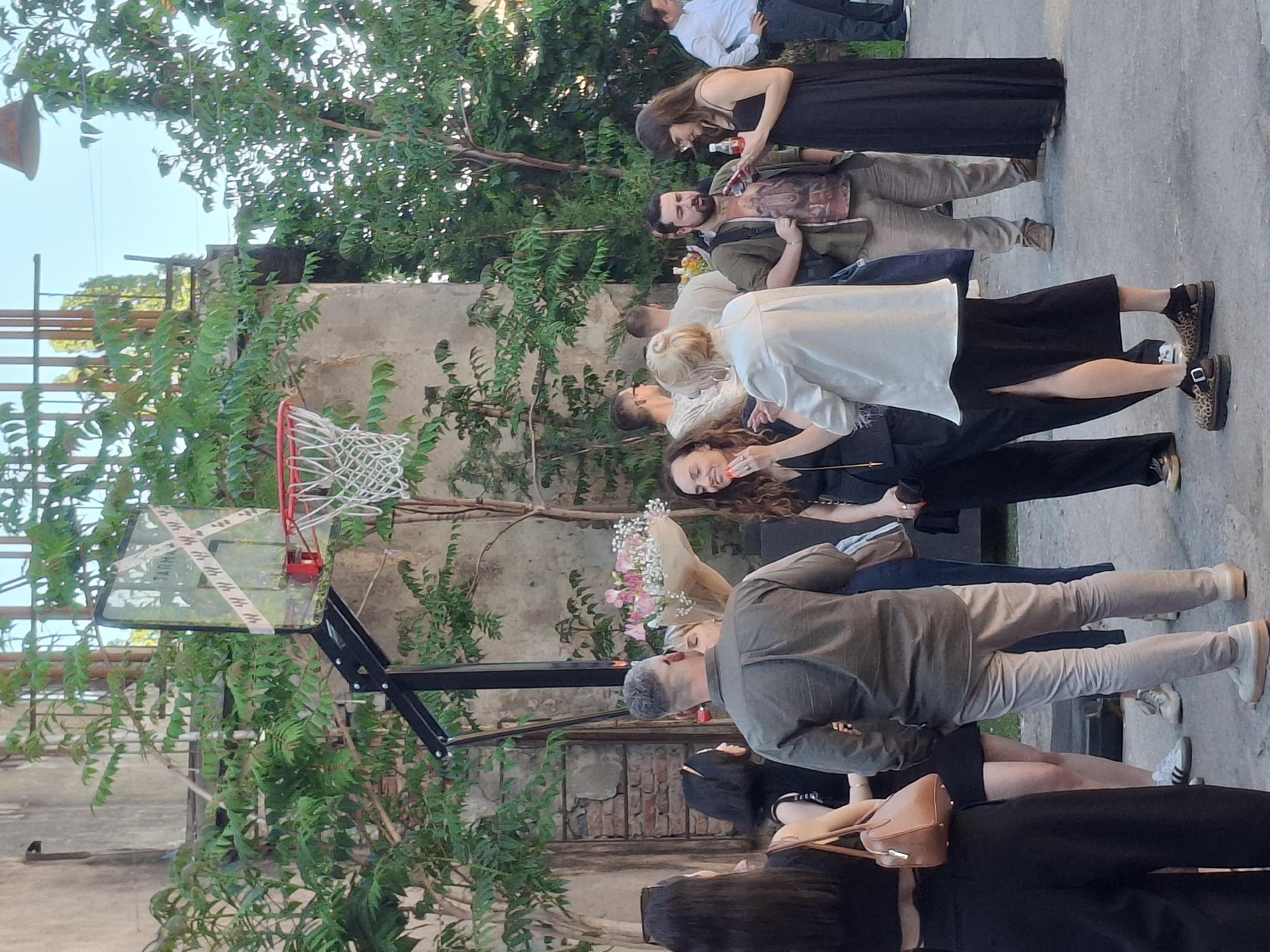 The image shows a gathering of people outdoors, possibly at a celebration or event. A basketball hoop stands prominently in the foreground, partially obscured by greenery. A group of people is seen standing and socializing, with their attire suggesting a semi-formal occasion. Several individuals are holding drinks, while another carries a bouquet of flowers. The background includes a brick wall and foliage, suggesting an urban environment, possibly a courtyard or garden. The overall lighting indicates it is daytime.