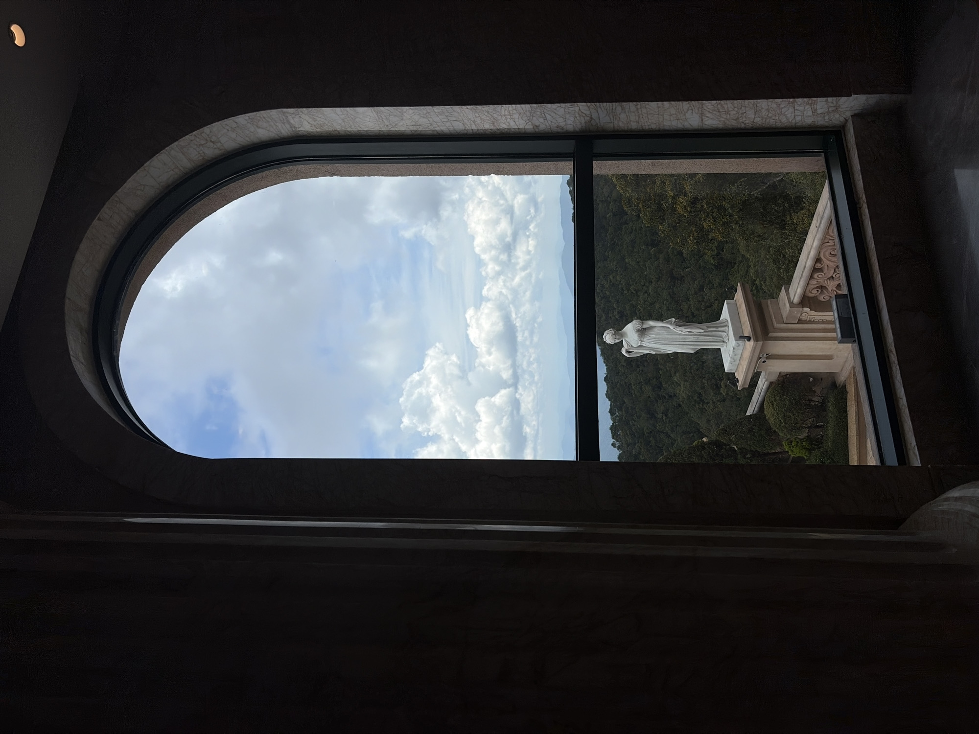 A view from inside a building, looking out through a combination of a curved and rectangular window. Outside, there is a statue of a woman standing on a pedestal, surrounded by trees and vegetation. The sky above is visible, filled with white clouds against a blue background. The interior of the building is dark, with marble or stone surfaces. The location is Bà Nà Hills, Đà Nẵng, Vietnam, specifically the Linh Ứng Pagoda.