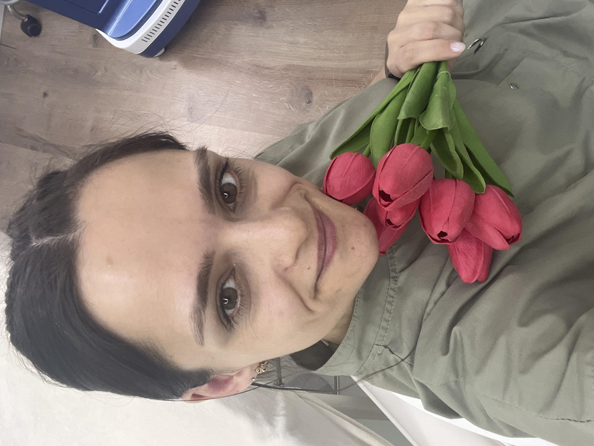 A medium shot captures a woman holding a bouquet of red tulips, likely indoors. The woman has dark hair and fair skin, and she is wearing a light green jacket. The location is suspected to be in Chaikovsky, Russia, based on contextual information. The environment includes what appears to be a wood-like surface and other indeterminate elements. The woman is looking directly at the camera.