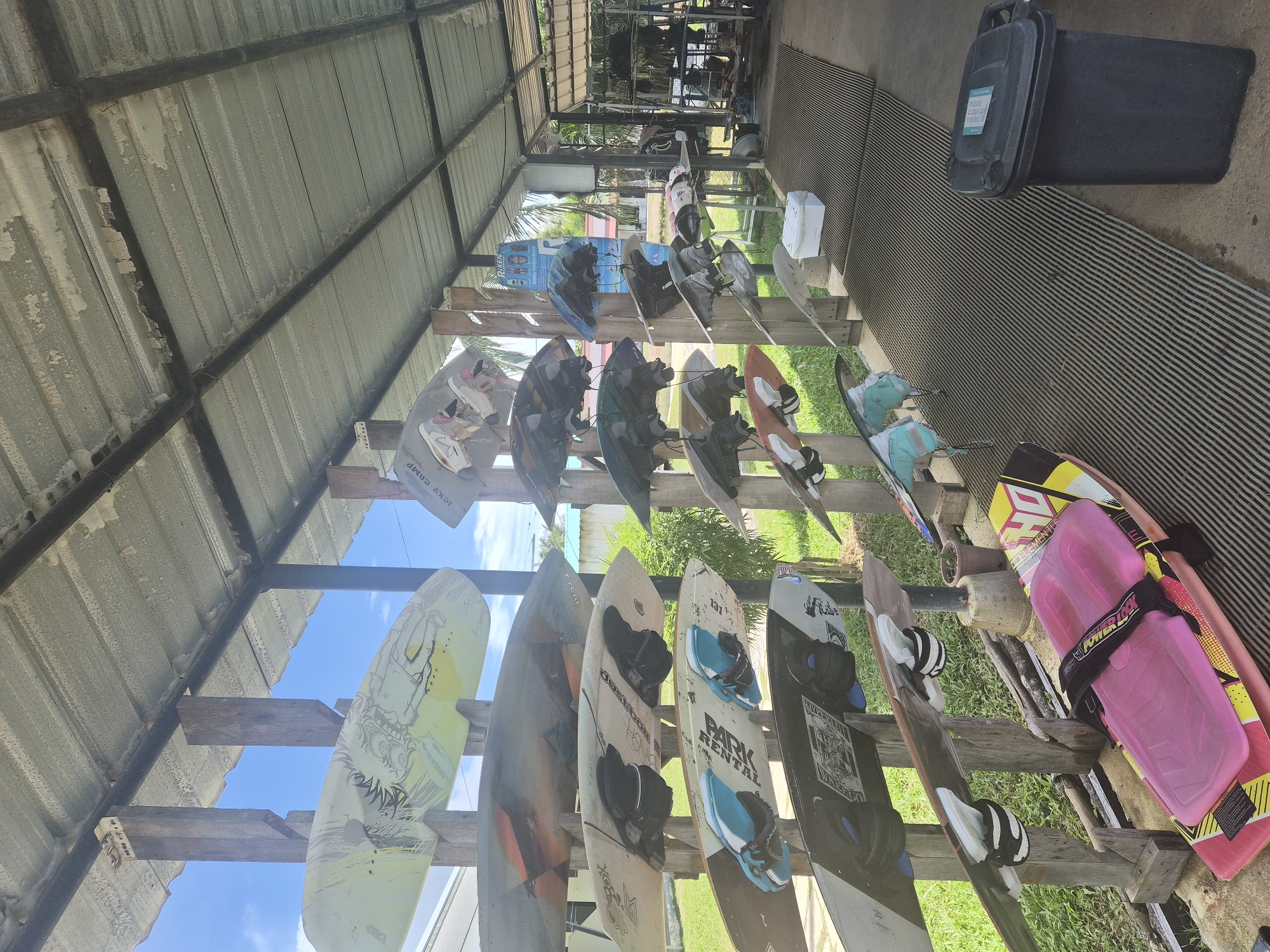 The image depicts an outdoor equipment storage area at a facility in Tambon Mai Khao, Thailand. The scene is dominated by a rack system holding numerous wakeboards and surfboards, oriented horizontally and vertically. The boards exhibit various colors and designs, with some featuring bindings.
The storage area is covered by a corrugated metal roof supported by wooden beams and metal poles. The roof shows signs of wear and peeling paint. Beneath the roof, a patterned rubber matting covers the ground.
To the left of the rack, lush green grass and palm trees are visible, suggesting a tropical environment. In the background, a white building with a light-colored roof can be seen, along with more palm trees.
On the right side of the image, a black trash bin with a light blue label is positioned on the matting. Further back, a metal rack holds more equipment, and a small white cooler sits on the matting.
There are no people visible in the image, and the focus is on the organized display of water sports equipment under the covered structure. The overall impression is of a functional, open-air storage facility for recreational watercraft.