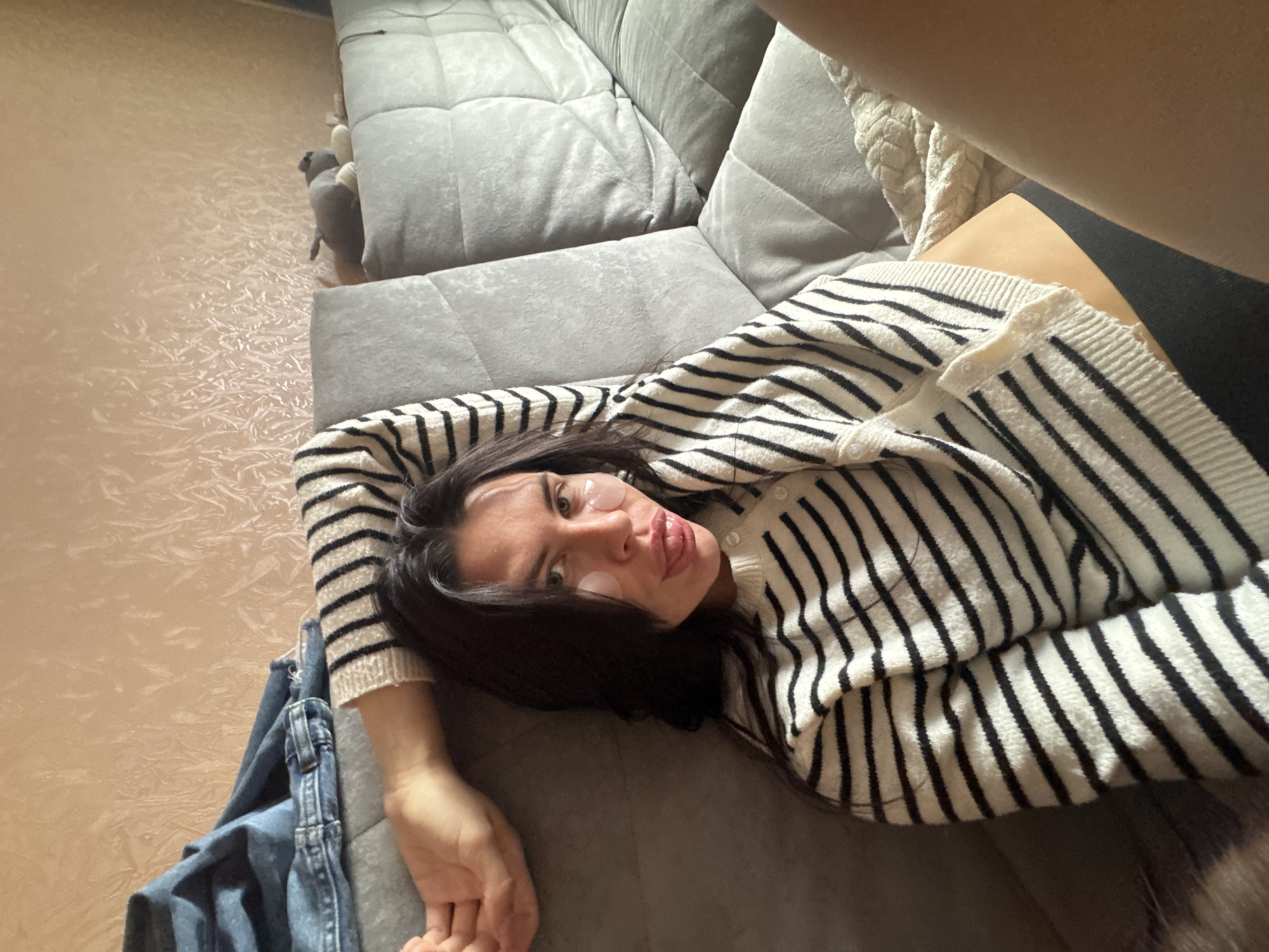 A woman with long dark hair is lying on a grey couch, wearing a black and white striped sweater. She has eye patches on her under-eye area and is sticking her tongue out. To her left, on the couch, are a pair of blue jeans. The background is a textured, light-colored wall. The couch appears to be in a residential setting, likely an apartment or house in Chaikovsky, Russia.