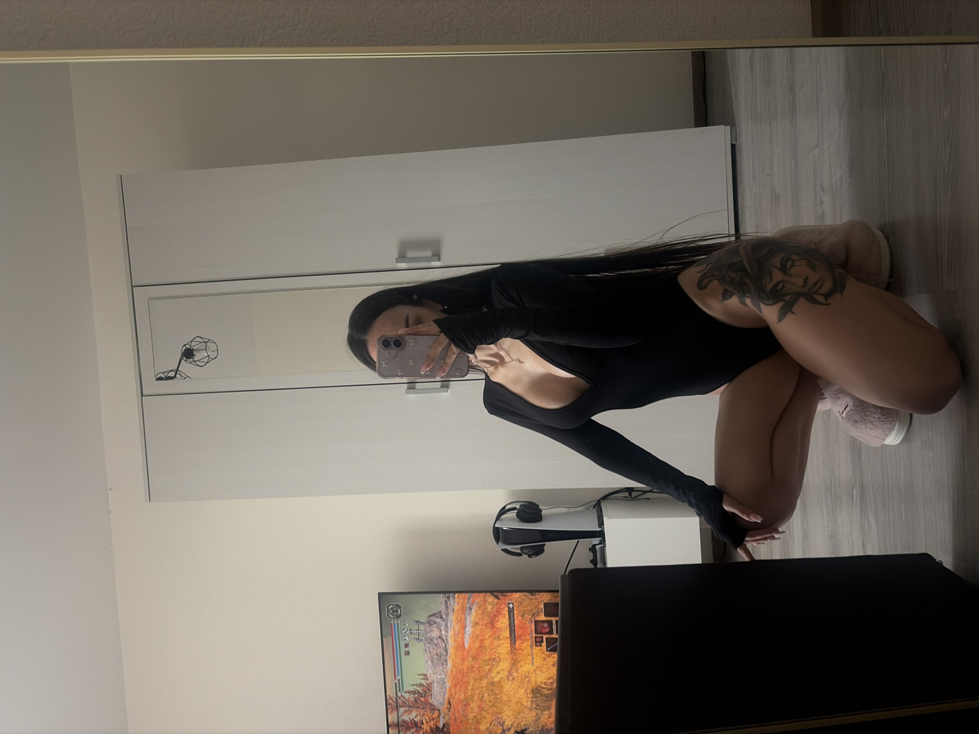 A female with long dark hair is kneeling in front of a mirror, taking a picture of herself with a mobile phone. She is wearing a black bodysuit. The background includes a white wardrobe, a television screen displaying a game, a PlayStation 5 console, and headphones. The flooring appears to be light grey. The location cannot be determined to be Saint Petersburg, Russia based on the information provided.
