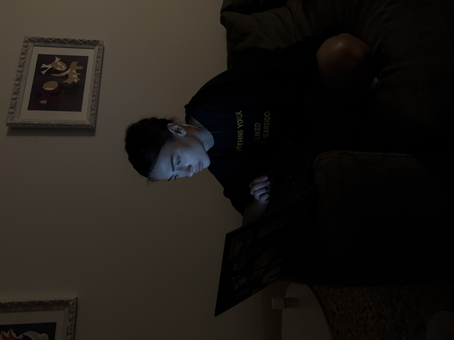 An indoor scene in Venice, Italy, depicts a lone female seated on a dark upholstered couch in a dimly lit room. The primary light source emanates from an open laptop computer positioned on her lap, casting a distinct bluish glow onto her face, neck, and hands. The individual has dark hair pulled back and wears a dark t-shirt, on which partially visible yellow-green text reads "VETHING YOULK" and "LIKED" followed by obscured characters. A white wired earphone is inserted into her right ear, and a small stud earring is visible. Her left hand rests near the laptop's keyboard/trackpad area. The wall behind her is a light, neutral color, adorned with two ornate white-framed artworks. The left frame contains an abstract piece with blue, red, and white elements, while the right frame holds an abstract composition featuring yellow, purple, and orange shapes. To the left of the couch, a partial view of a round white side table is visible, upon which a white cylindrical container, possibly a pill bottle with an illegible label, rests. The setting suggests a late-night or low-light environment.