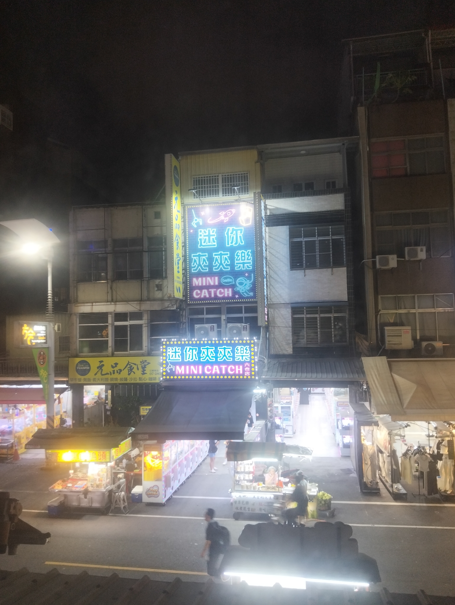 The image depicts a night scene on a street in Kaohsiung, Taiwan, likely in an urban commercial district. The foreground shows a street with vehicles and pedestrians, blurred due to movement. In the midground, several street vendors are operating. One vendor appears to be selling food from a cart illuminated by bright yellow lights, with products displayed on shelves. Another vendor has a row of vending machines. In the background, buildings line the street, featuring illuminated signs with Chinese characters and English text. One prominent sign reads "MINI CATCH" in neon lights, along with Chinese characters and cartoonish illustrations of a rocket and an astronaut. The architecture suggests multi-story buildings with visible windows and air conditioning units. The overall atmosphere is typical of a bustling night market or shopping street.