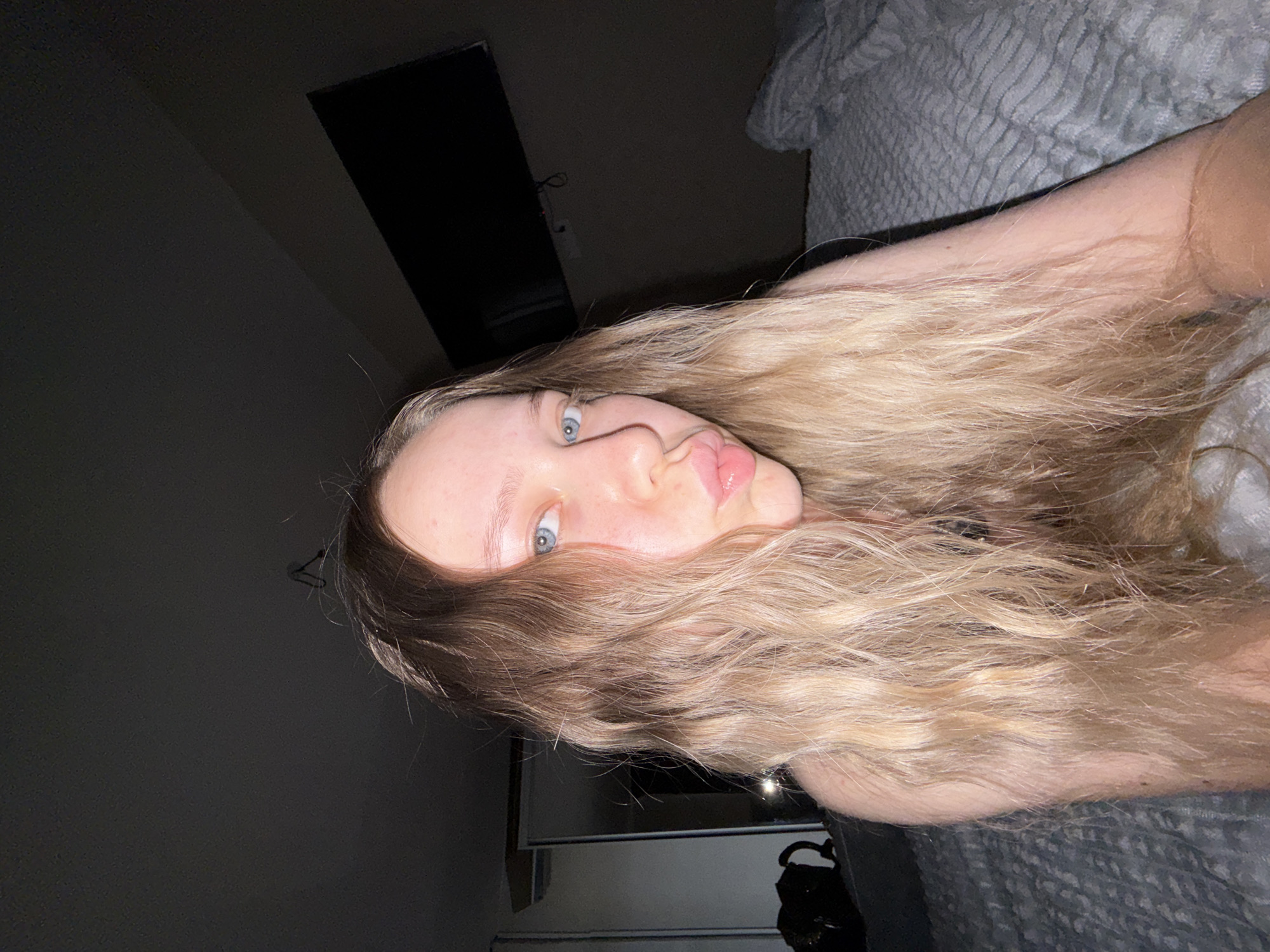 A young woman with long blonde hair and blue eyes is lying down in a bed with gray patterned bedding. She is positioned facing upwards, with her head tilted to the left. The background includes a television mounted on the wall and a dimly lit corner of the room. The overall setting appears to be a bedroom. There is no evidence of this image being from the city of Chaikovsky, Russia.