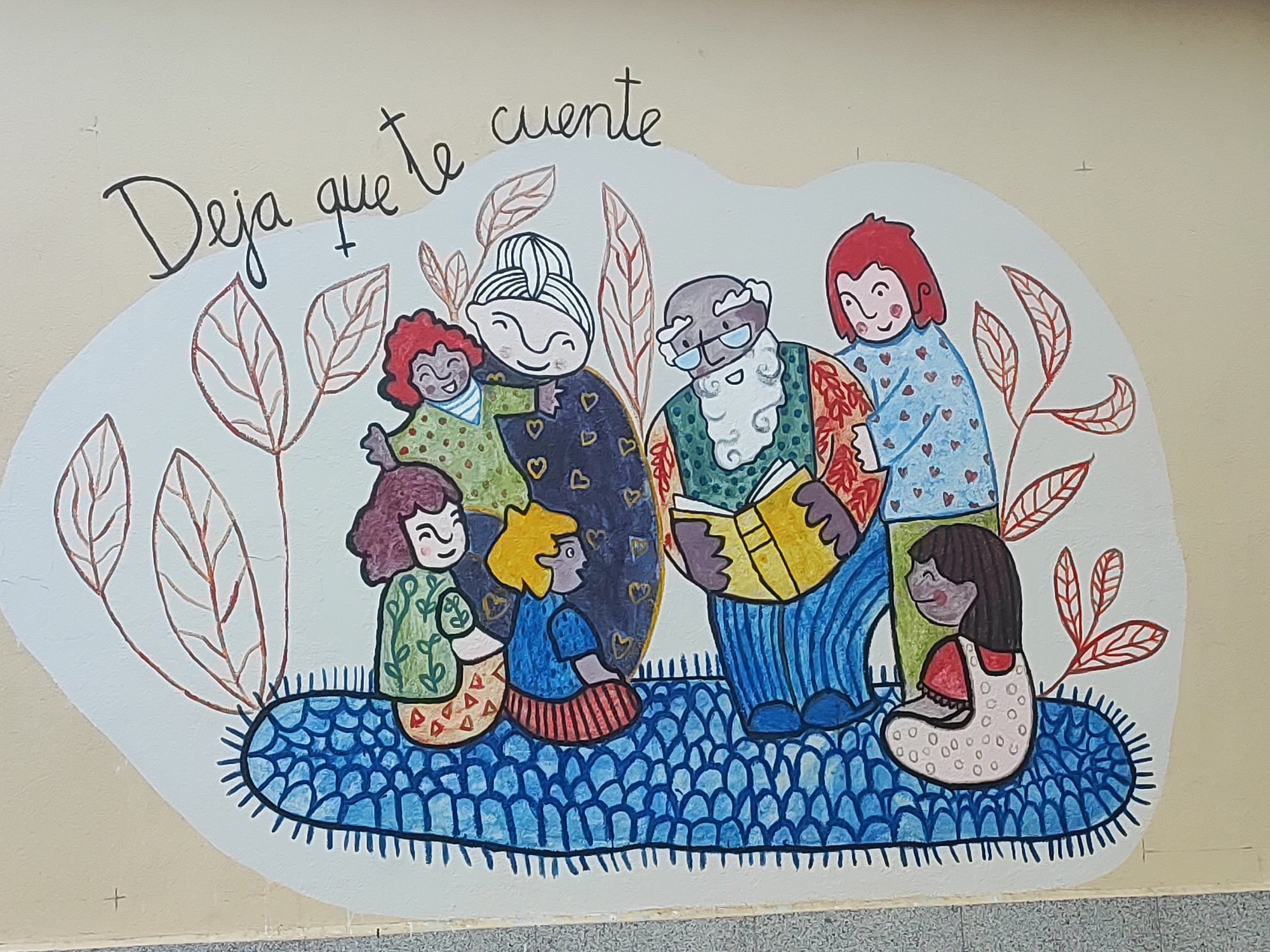 This is a colorful mural painted on a light-colored wall, likely outdoors or in a public space in Torre De Don Miguel, Spain. The mural depicts a group of people of various ages gathered together, seated on a blue, textured mat. The overall theme appears to be storytelling or reading.
In the foreground, a young girl with dark hair, wearing a light-colored dress with pink polka dots and a red undershirt, is seated on the mat with her legs tucked. To her left, another child with blonde hair and a blue shirt is also seated, looking towards the center of the group.
In the center of the mural, a man with glasses, a white beard, and a mustache is reading a yellow book. He is dressed in a blue striped outfit and a patterned vest. Next to him, an older woman with grey hair tied up, wearing a dark blue dress adorned with hearts, has her eyes closed and a serene smile. To her left, a young child with red curly hair and a green polka-dot dress is laughing. On the far right, a child with short red hair and a blue shirt decorated with small red hearts is smiling.
Surrounding the group are stylized drawings of brown leaves with red veins, adding to a natural or autumnal theme. The mural is bordered by a white outline, and the Spanish phrase "Deja que te cuente" (Let me tell you) is written in black cursive script above the figures. The bottom of the mural is close to a textured gray surface, possibly a sidewalk or base of the building.