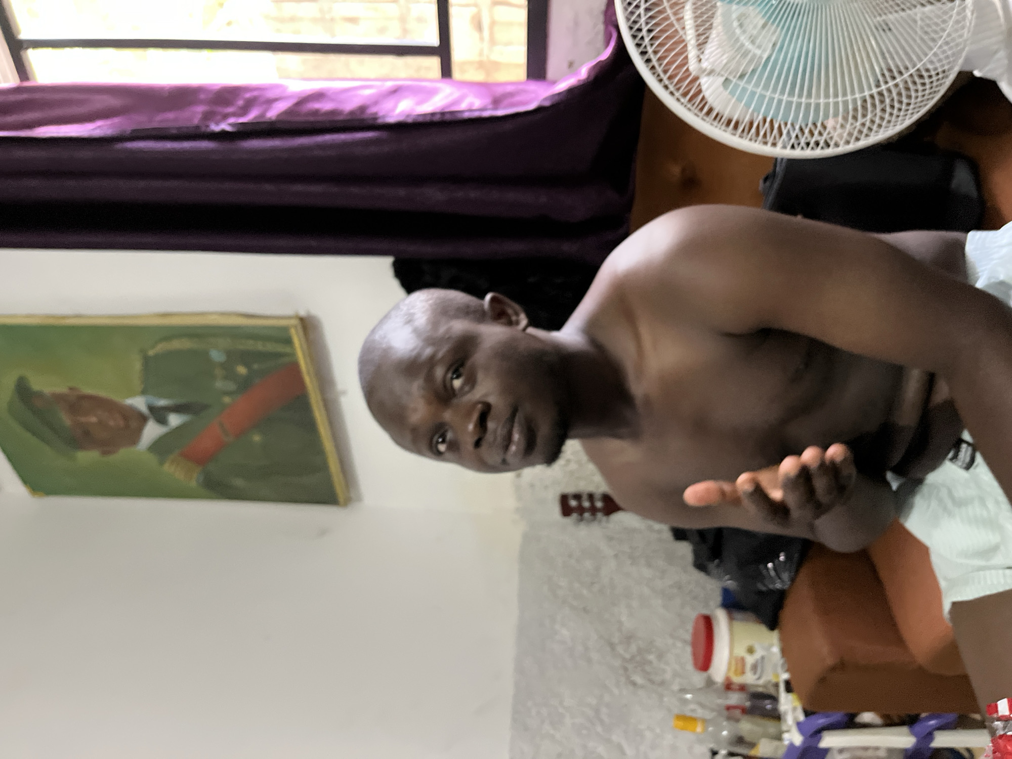 A medium-angle shot shows a shirtless dark-skinned man with a bald head. He is looking at the camera, with his right arm extended towards the viewer. Behind him is a room with a window draped with purple fabric. To the left is a framed painting of a person in military uniform. In the background, there is a white fan and some cluttered items on a surface below, including bottles and a red-lidded container. The exact location in Abuja, Nigeria, cannot be determined from the image.