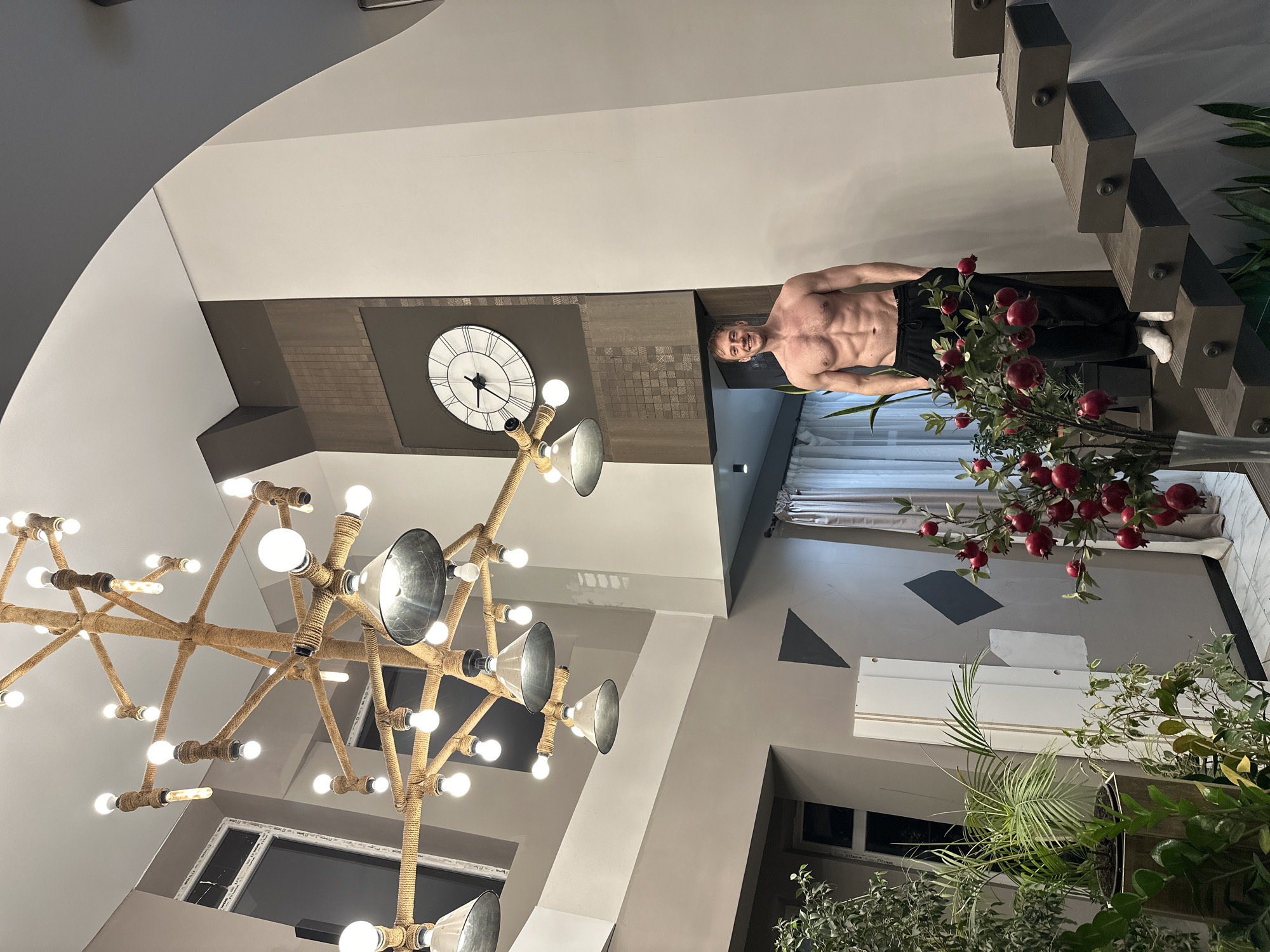 A sparsely furnished interior is viewed from a low-angle perspective, dominated by a decorative chandelier in the foreground. A shirtless man stands adjacent to a modern staircase. To the left of the staircase stands a decorative branch. A clock is mounted on the wall. The exact location in the city of Kursk, Russia, cannot be determined from this image.