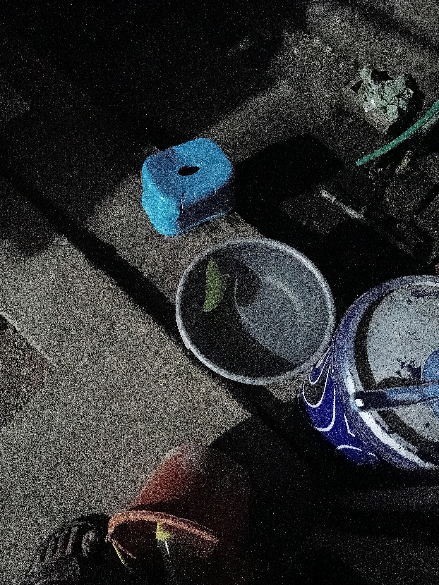 In Cikal, Indonesia, an outdoor scene is depicted, likely a backyard or alleyway, illuminated by a single light source casting a long shadow from a blue stool. The ground is composed of rough, textured concrete.
Several objects are present:
* A bright blue, cubical plastic stool with a circular opening in its top is positioned towards the upper left of the frame, casting a shadow across the ground.
* To the right of the stool, a gray plastic basin is partially visible, containing a single green object, possibly a vegetable or a leaf.
* To the right of the basin, a blue and white patterned bucket is seen, with a white lid partially obscuring its contents.
* In the foreground, at the bottom left, a portion of an orange bucket or container is visible, with what appears to be a black shoe or boot placed on top of it.
* Further back on the right side, there is a pile of green debris next to a portion of a green hose. A metal pipe is also visible in this area.
The overall impression is one of a functional outdoor space, possibly used for domestic chores or storage, given the presence of buckets, a stool, and a hose. The lighting suggests it is either early morning or late evening.