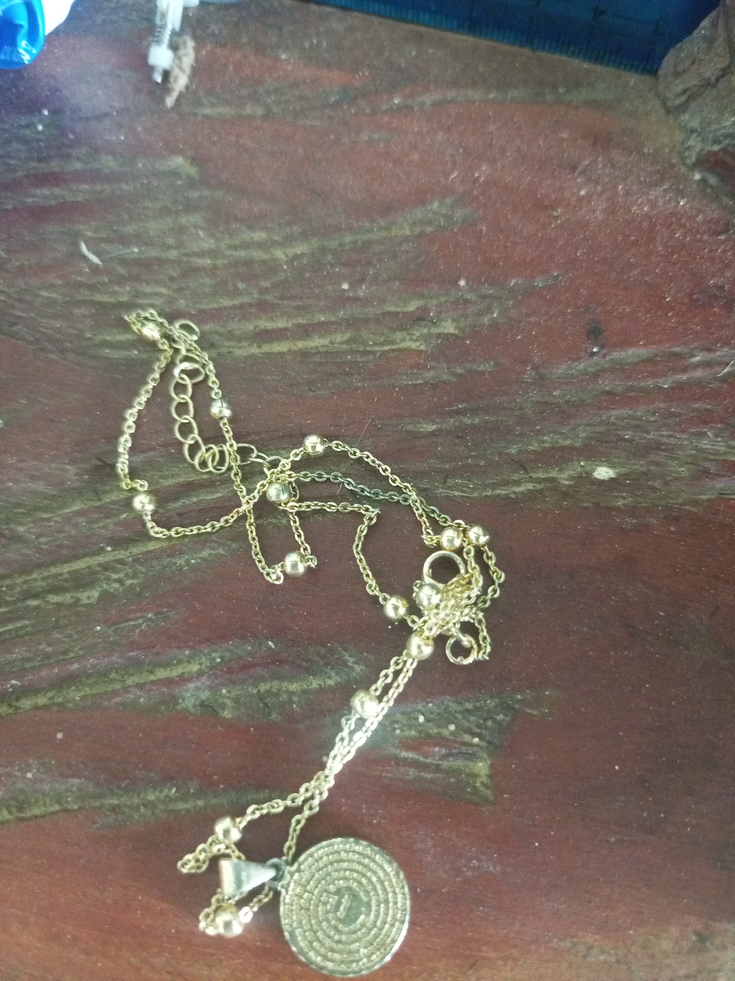 The image shows a close-up, overhead view of a gold-colored necklace with small beaded accents and a circular pendant resting on a dark, textured wooden surface. The necklace is draped across the surface in a loose, meandering pattern, with the pendant positioned in the lower right quadrant of the frame. The wooden surface appears to be aged and slightly weathered, with visible grain and some scattered debris or dust. The background is out of focus, but suggests a rustic or natural environment. There is no indication of the location being within the city of La Tagua, Venezuela, or any other specific geographical location. The image is static and shows only inanimate objects.