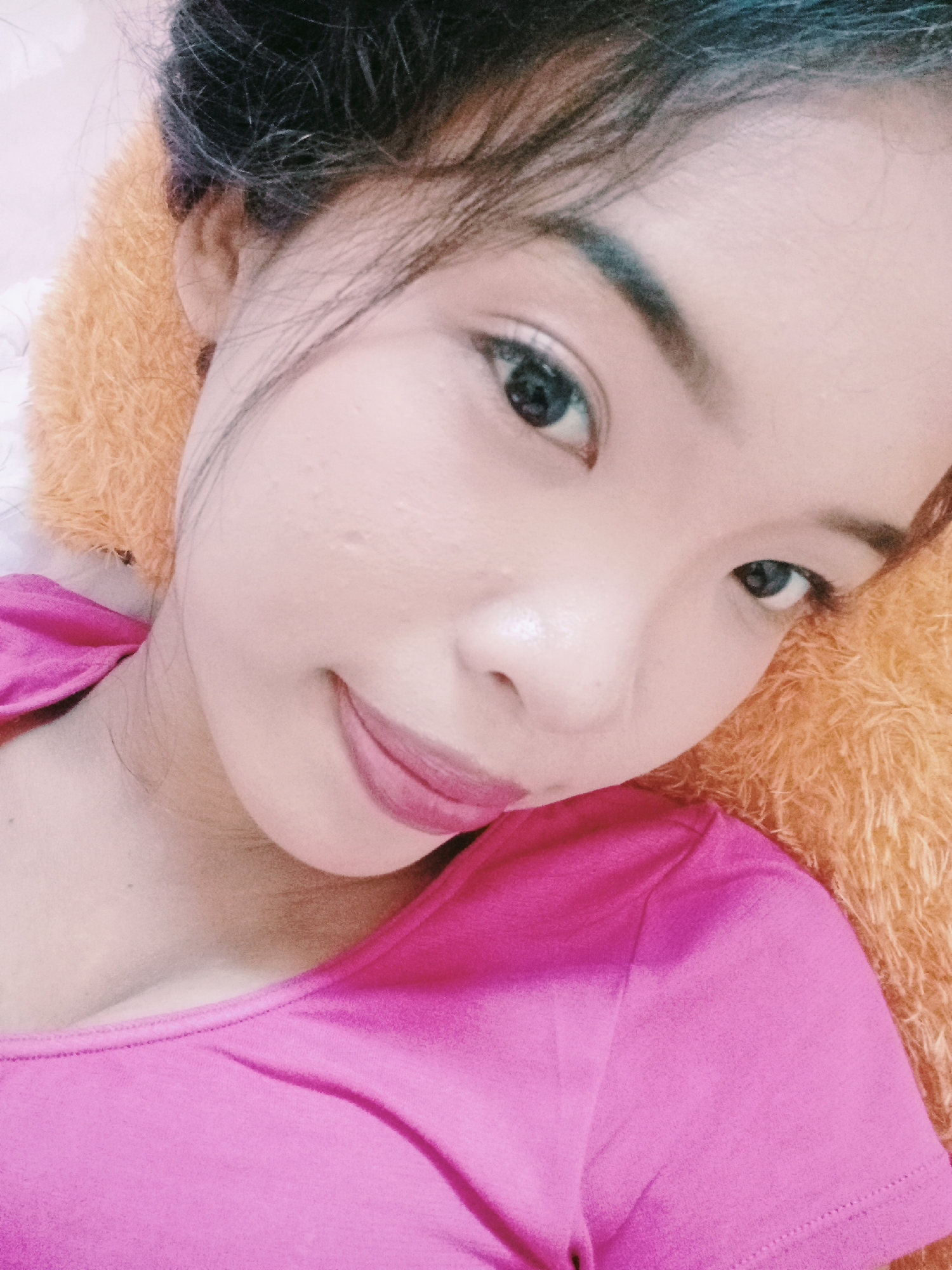 The image is a close-up, over-the-shoulder selfie of a young woman, likely in a relaxed, indoor setting. She is lying down, with her head resting on an orange, fluffy surface that appears to be a pillow or blanket. Her face is turned towards the camera, and she is looking directly at it. She has dark hair pulled back, and is wearing a bright pink top. Her makeup includes dark eyebrows and pink lipstick. The background is softly blurred, suggesting a shallow depth of field. The overall lighting is soft and warm. There is no indication of the city of Pamesi, Indonesia in the image.