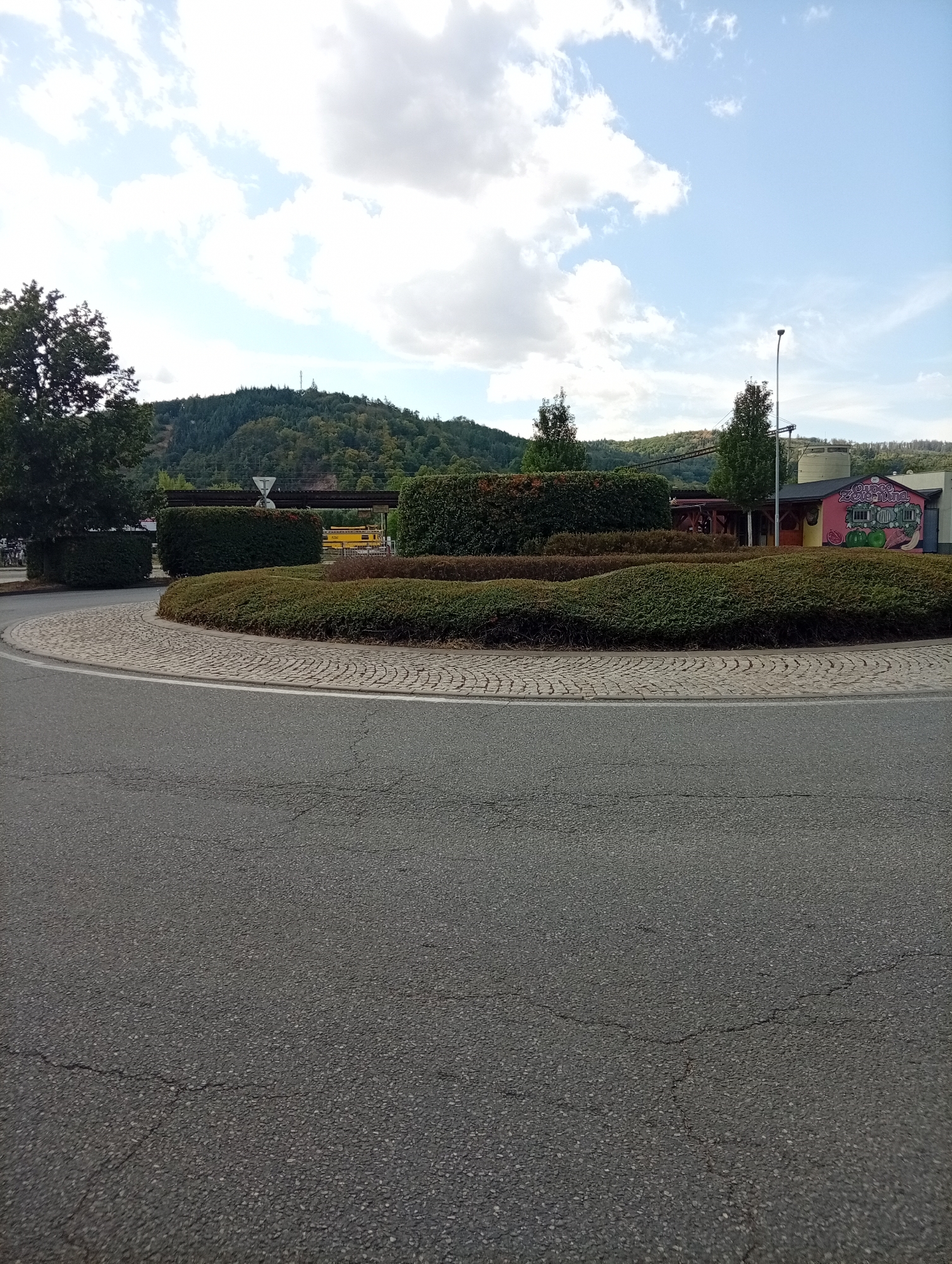 The image shows a roundabout with a circular flower bed in the center. The roundabout is paved with cobblestones and surrounded by asphalt road. Behind the flower bed, there is a building with a colorful mural on its side, and a tall industrial structure. The background consists of a forested hill. This location appears to be on the outskirts of Tišnov, Czechia, possibly near an industrial area or a recreational facility. The sky is partly cloudy. No people are visible in the image.