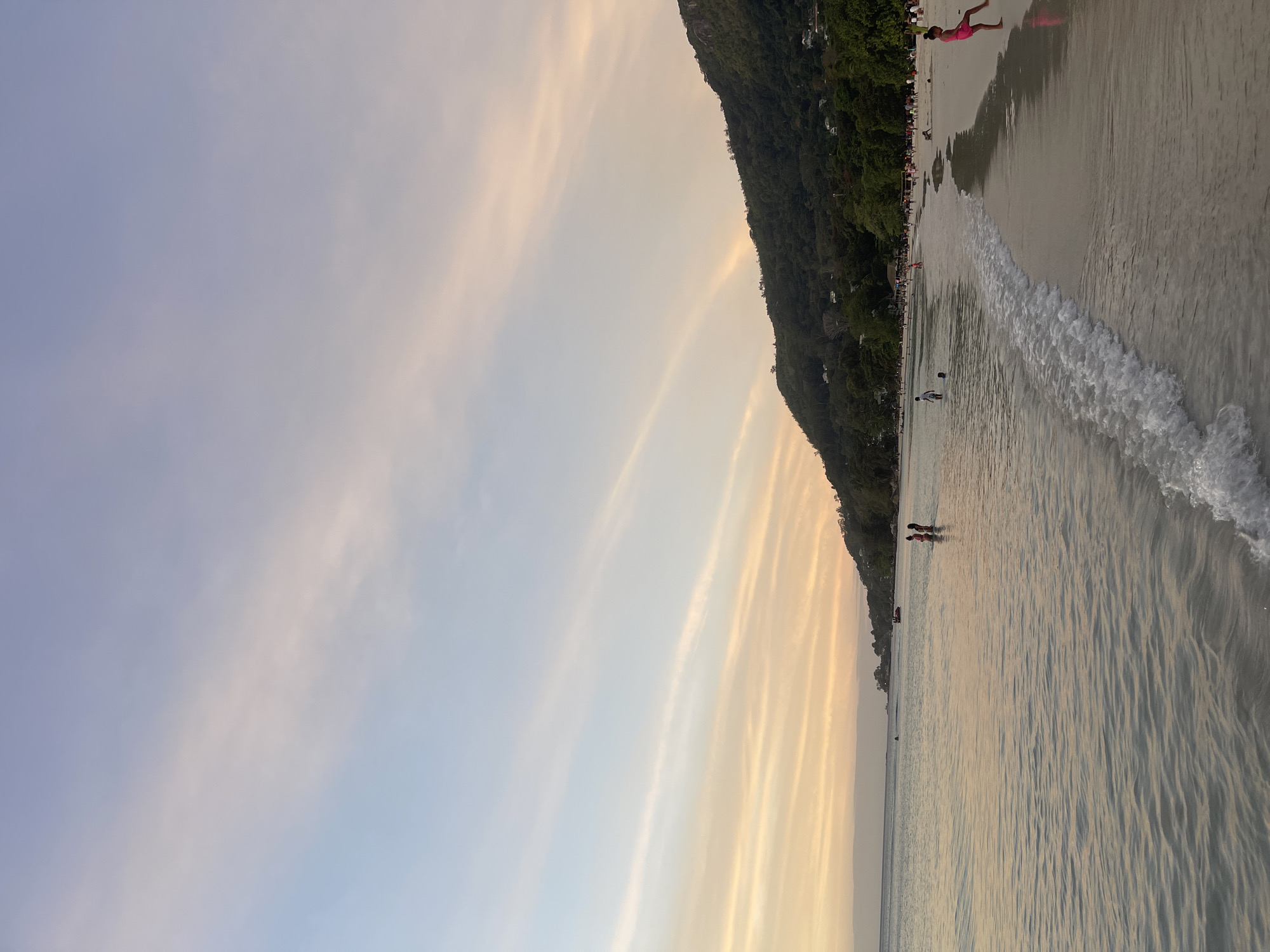 This image depicts a beach scene at Mare Anglaise, Seychelles, during what appears to be sunset or sunrise. The sky is a gradient of pale blue and soft orange hues, with thin, elongated clouds streaking horizontally. A lush, green, mountainous coastline forms the backdrop on the right side of the frame, dotted with some houses. The foreground is dominated by a calm, shallow body of water reflecting the sky's colors, with gentle waves lapping at a sandy beach. Several people are visible: a few individuals are wading in the water, while others are on the beach, some appearing to be playing or walking. A jet ski is visible in the distance on the water. The overall atmosphere is tranquil and serene.
