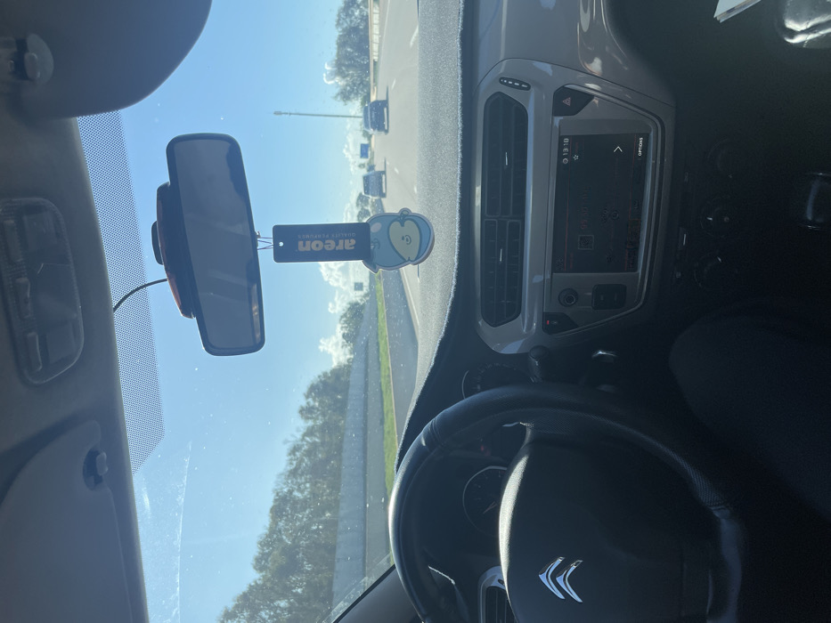The image depicts an interior view from a Citroën vehicle, positioned as if from the driver's seat, looking forward onto a multi-lane highway near Skhirat, Morocco, during daylight hours. A driver's left arm and leg, clad in dark clothing, are partially visible. The black steering wheel, featuring a silver Citroën emblem, is prominent on the left. The grey dashboard extends across the foreground, partially covered by a dark, textured mat. A central infotainment system displays the time as "13:18" and "Radio" mode, flanked by air vents and climate control knobs. A rearview mirror is mounted centrally above the dashboard, with a small electronic device attached above it. Suspended from the rearview mirror is a blue, cartoon-character shaped air freshener bearing the "Areon" brand.
Through the windshield, the highway, marked by clear asphalt, stretches into the distance, with three other vehicles visible ahead, traveling in the same direction. To the left of the highway, a concrete barrier separates the road from a dense line of green trees. To the right, a wide shoulder or median leads to additional vegetation and a tall, slender pole. The sky above is clear blue with scattered white cumulus clouds, indicating bright, sunny weather.