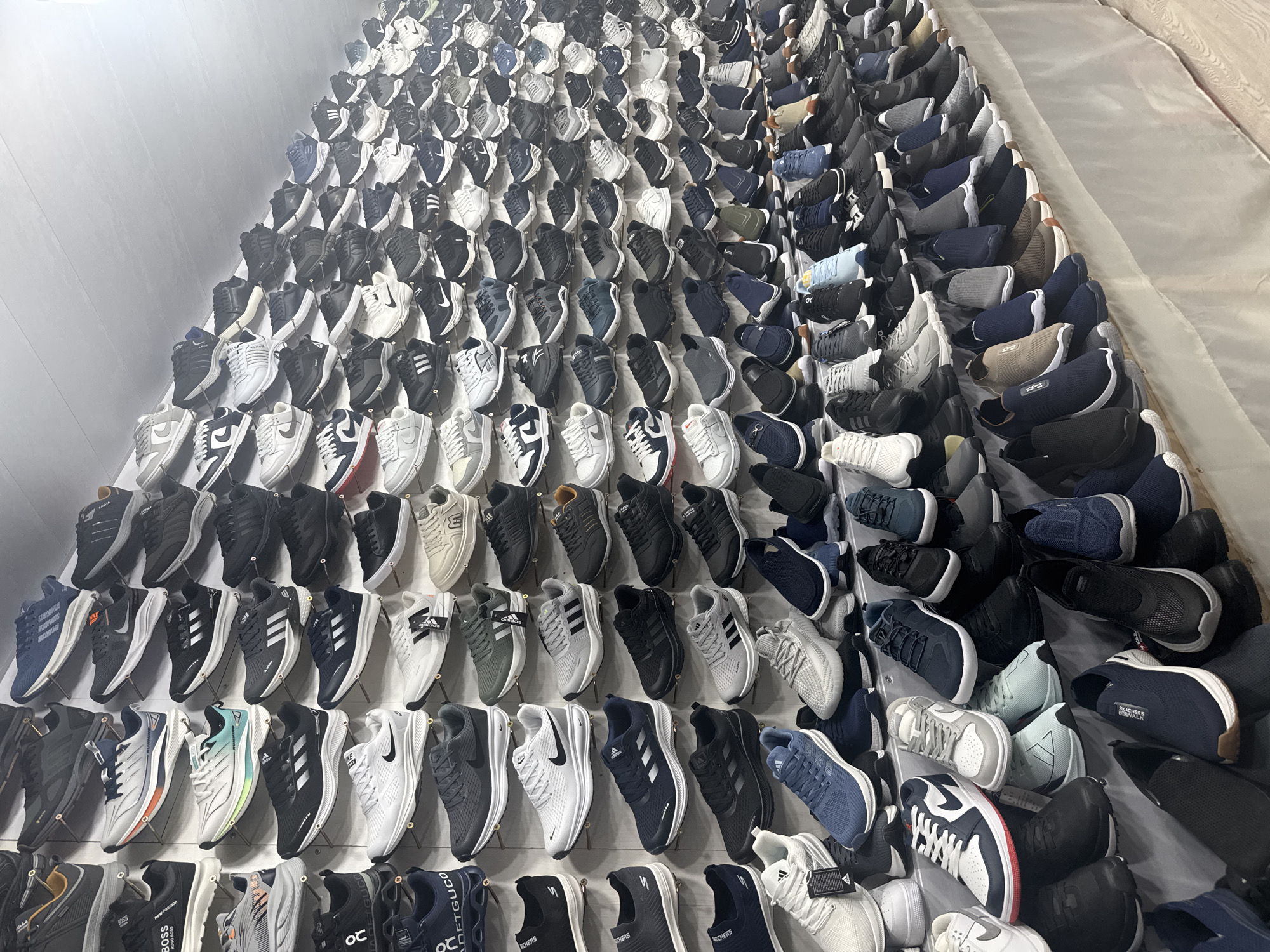 A large wall display of hundreds of athletic shoes is the central focus of the scene. The shoes are arranged in an organized manner, mostly in rows and columns, creating a wall of footwear. The shoes are mostly black, white, and grey. The environment appears to be a retail or storage space. This scene is located in Darkhan, Uzbekistan.