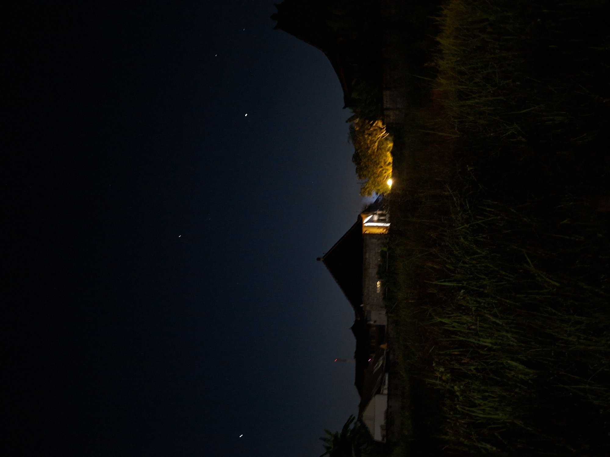 The image depicts a nighttime scene in the Ubud District of Indonesia. The sky is dark, with a few visible stars. In the foreground, there is a house with a sharply angled roof. A light shines from a tree near the house. To the right of the house, there is tall grass. Other buildings and plantlife can be seen but are obscured by darkness.