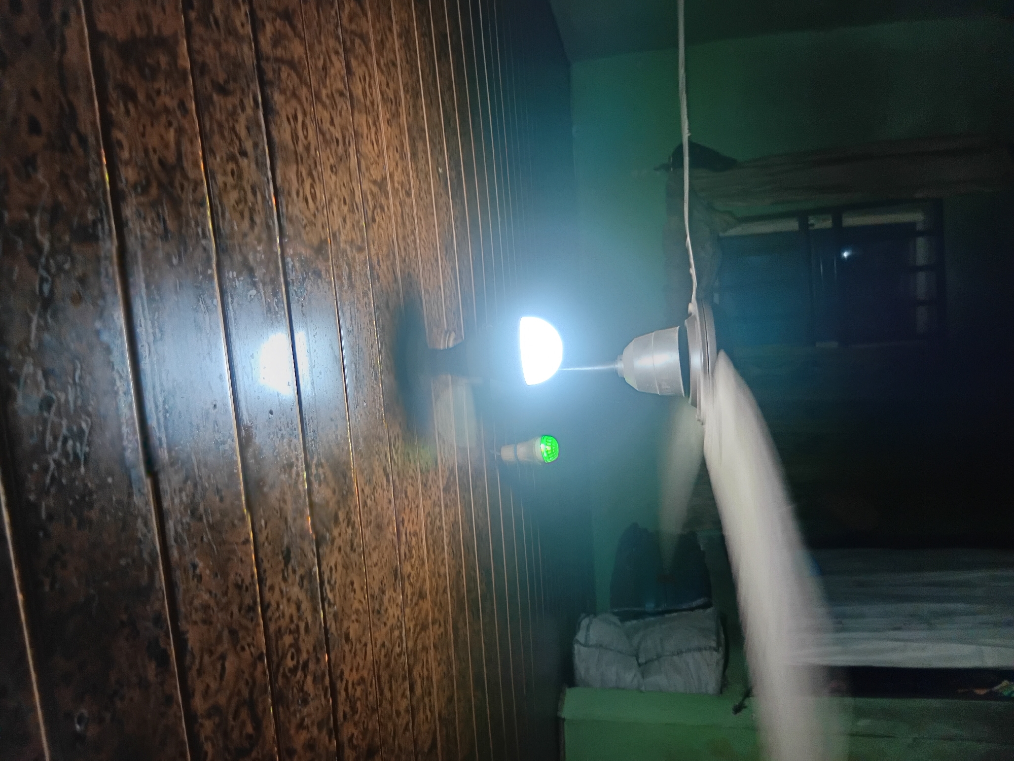 The image depicts the interior of a room with a textured, dark brown wooden slatted ceiling. Two light bulbs are visible; one is a bright white LED bulb suspended from the center of the room, and another smaller, green LED bulb hangs to its left. Below the lights, a ceiling fan with silver blades is in motion, creating a blur effect on the blades and the air around them. To the left of the fan, a stack of large, white bags or sacks is visible against a green wall. A window with bars is partially obscured by curtains on the right side of the frame. The overall scene suggests a simple, possibly residential or storage space in Nkpor, Nigeria, illuminated by artificial lighting.