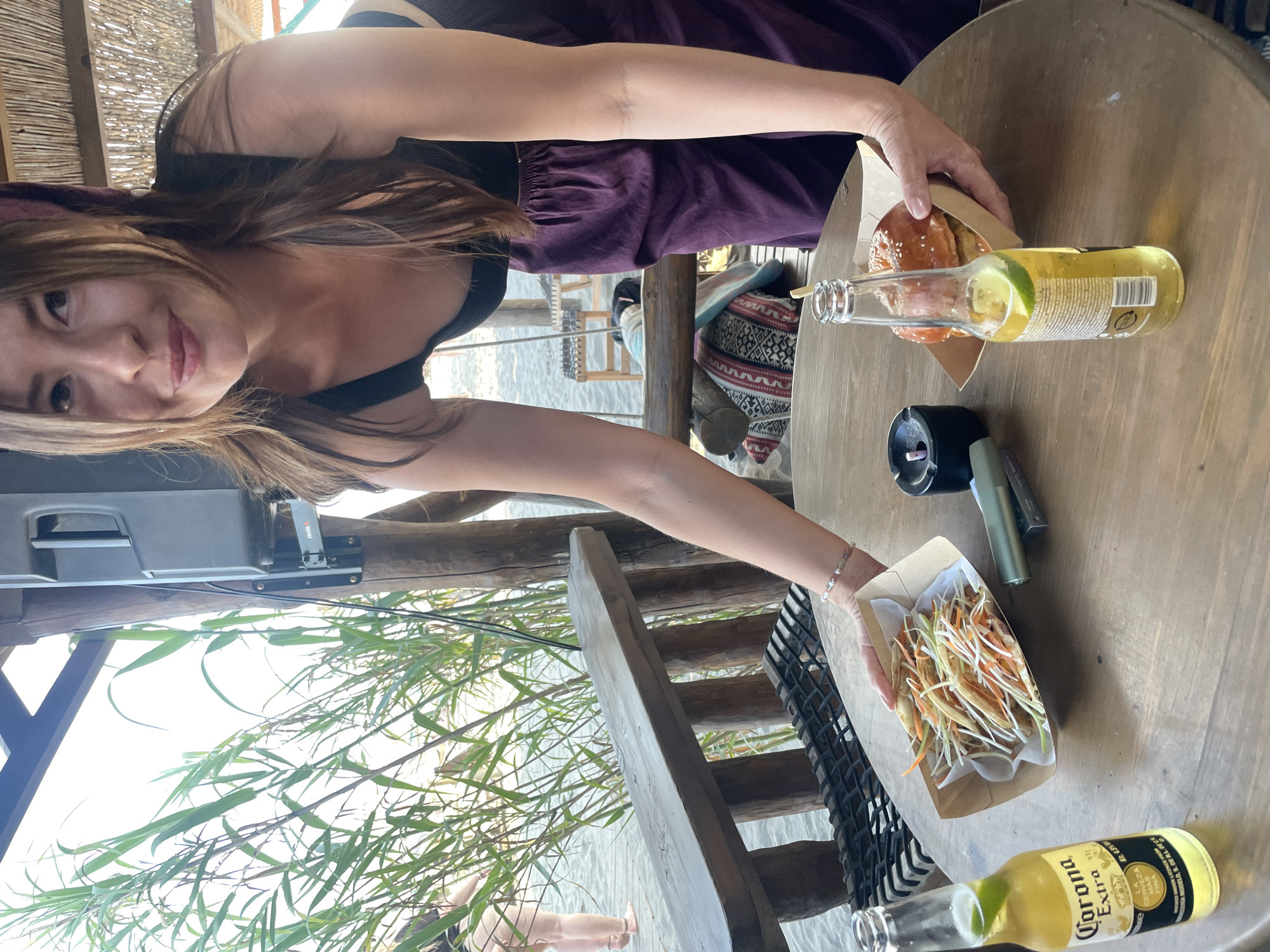 In Donji Štoj, Montenegro, a woman with long brown hair, wearing a black top and a purple skirt, is leaning forward to place a dish of food on a round wooden table. The table also holds a bottle of Corona Extra beer with a lime wedge, a burger in a cardboard container, an ashtray with a cigarette butt, and a green object resembling a lighter. The background shows a beach setting with sandy ground, wooden structures, and tall green plants. Another person is partially visible in the far left background. The scene appears to be outdoors, possibly at a beachside restaurant or bar.