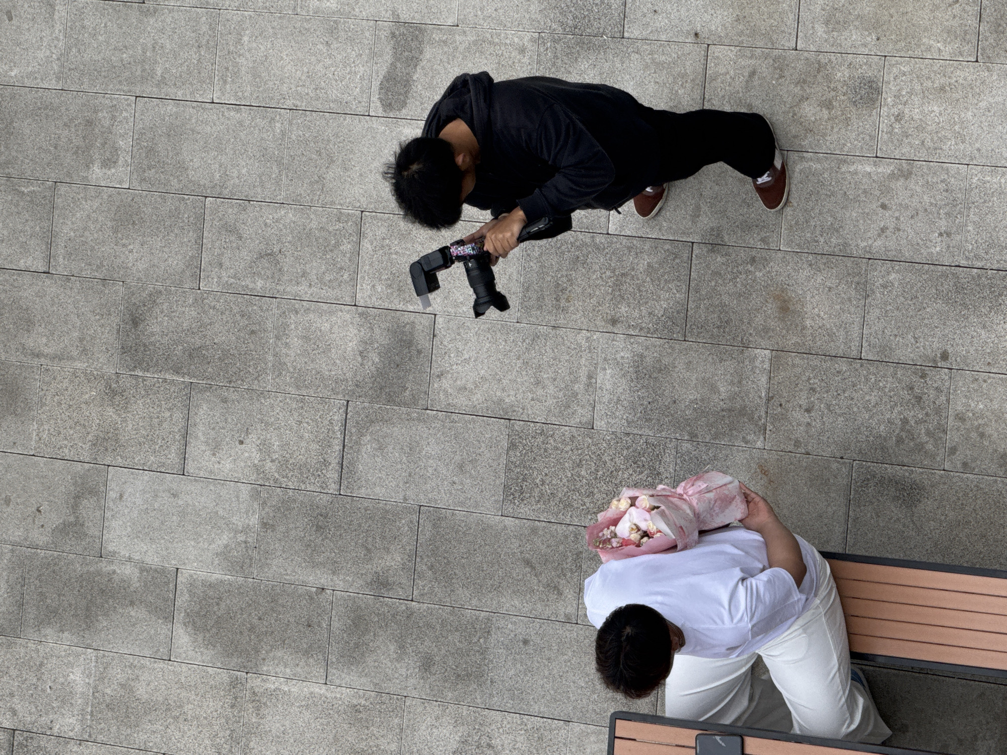 The image is taken from above and shows two people on a gray, brick-paved area in Zhongtun, China. One person is wearing a black hoodie, black pants, and brown sneakers. They are holding a camera with a flash attachment and appear to be taking a photograph. The second person is sitting on a wooden bench, wearing a white shirt and white pants, holding a bouquet of flowers wrapped in pink paper. A smartphone is on the bench near the person. The scene is outdoors, with the brick pavement filling much of the frame.