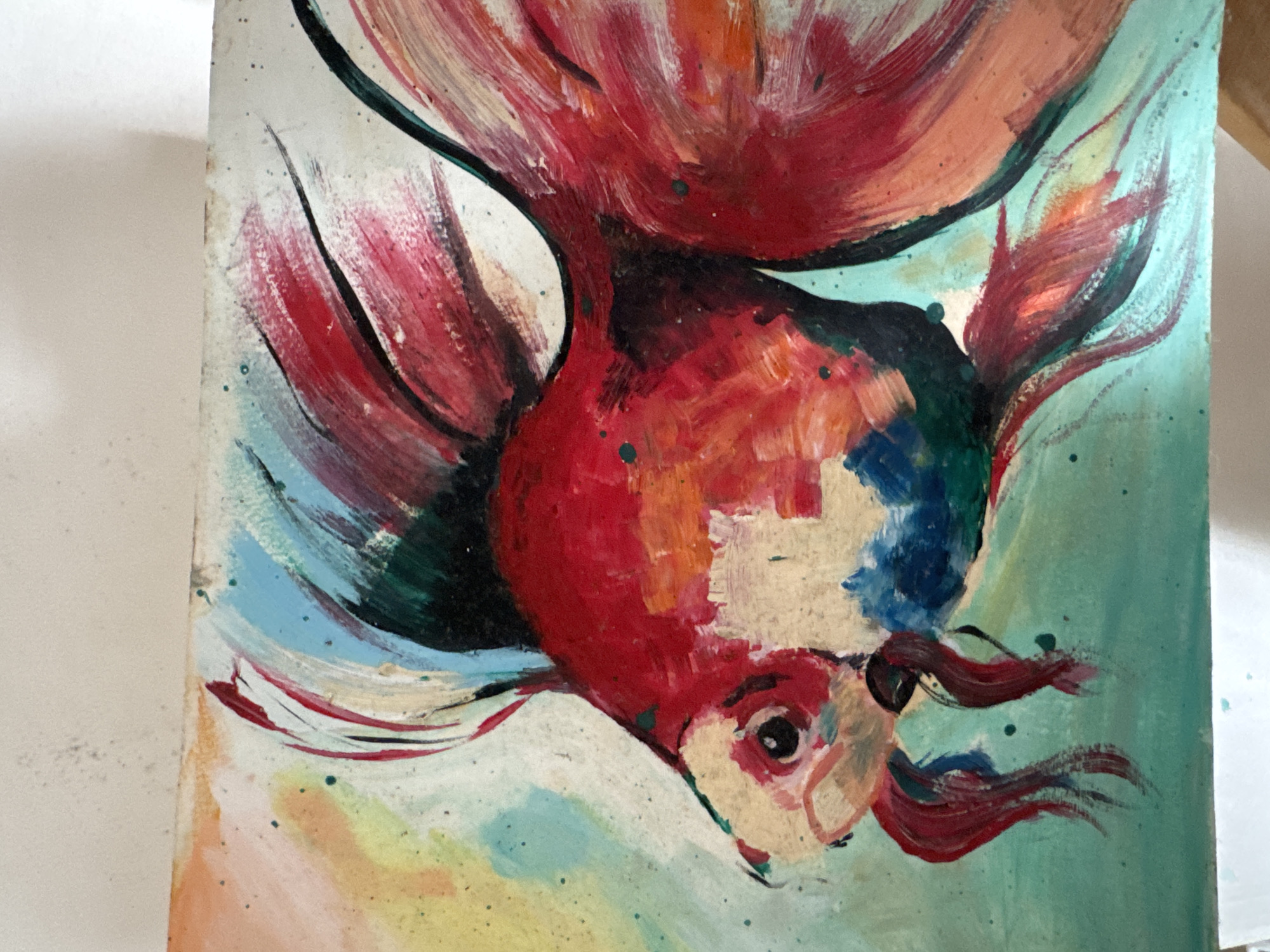 The image is a close-up, horizontal shot of a painting of a goldfish. The goldfish is rendered in a vibrant, impressionistic style with bold brushstrokes and a colorful palette. Dominant colors include shades of red, orange, white, blue, and green. The background is a wash of light colors, including yellow, orange, and light blue, with some speckles of dark paint. The painting appears to be on a canvas or textured paper. There is no indication of the location in Kuta Selatan, Indonesia, or any contextual information related to that specific place. The focus is solely on the artistic depiction of the fish.