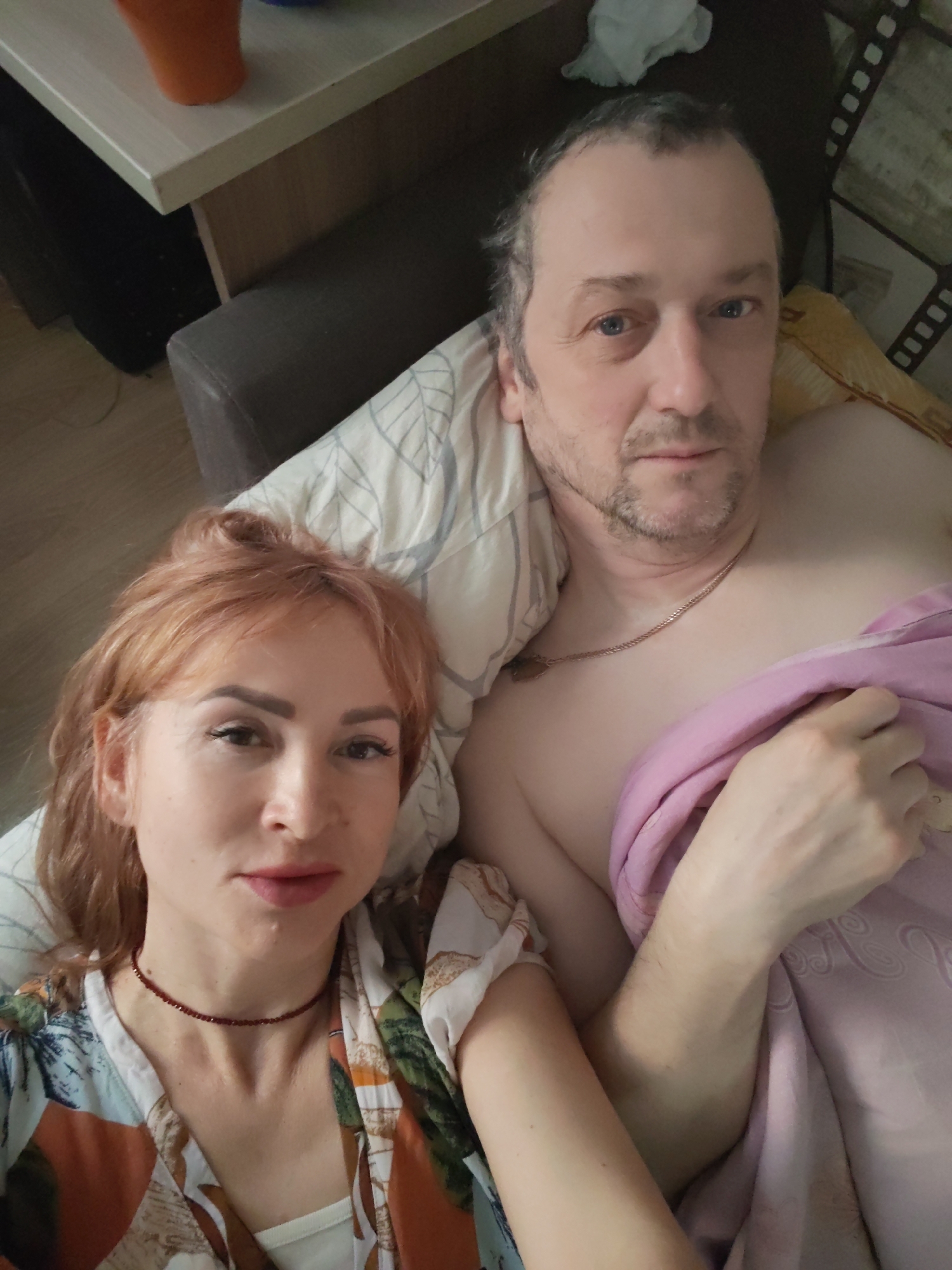 **Location:** The image appears to be taken indoors, possibly in a residential setting in the city of Chaikovsky, Russia. The presence of a blanket, bed linens, and a bedside table suggests this is a bedroom.
**People:** Two individuals are visible.
* A woman with reddish-blonde hair, fair skin, and a floral-patterned top. She is wearing a red choker necklace.
* A man with receding grey hair, fair skin, and a light beard. He is wearing a gold chain necklace and is covered by a pink blanket.
**Objects:**
* Bed linens: striped pillow, yellow blanket
* Pink blanket
* Bedside table with an orange object on top
* Choker necklace
* Gold chain necklace
**Actions/Interactions:** The two people are lying in what appears to be a bed, positioned close to each other, and looking at the camera.
**Environment:** The scene is dimly lit, which suggests the room may not have sufficient natural light. The overall environment appears to be casual.