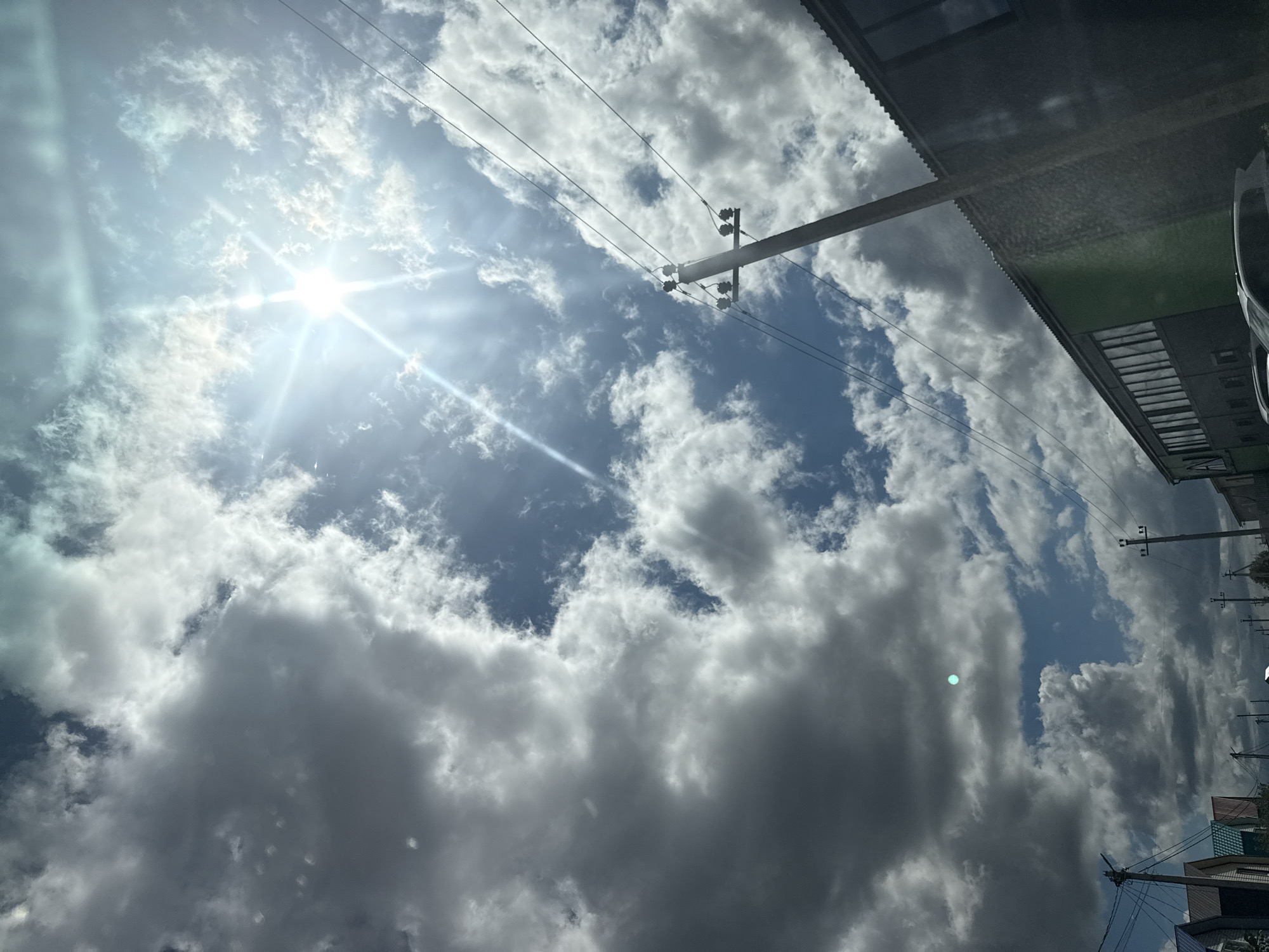 This is a photograph taken from a low-angle perspective, looking upwards at a dynamic sky filled with cumulus clouds. The sun is bright and appears to be high in the sky, with visible lens flares and rays of light streaming through gaps in the clouds. The sun's intensity suggests it is midday. The clouds range from bright white, where they are catching the sun's direct light, to dark gray, indicating depth and thickness.
In the foreground and mid-ground, several elements suggest an urban or semi-urban environment. A utility pole with insulators and wires is prominent on the right side of the image, stretching diagonally upwards. More power lines crisscross the sky. Partially visible on the right edge is the roofline of a building, appearing to have a corrugated metal surface and a green section. Further down the building, architectural details like windows and a roof are visible, with a hint of a car parked nearby. The overall scene is one of a bright, possibly warm day with a mix of sunshine and cloud cover. There is no visible text in the image. The angle and composition give a sense of looking up from street level, possibly from a car or a sidewalk, towards the sky and the overhead infrastructure.