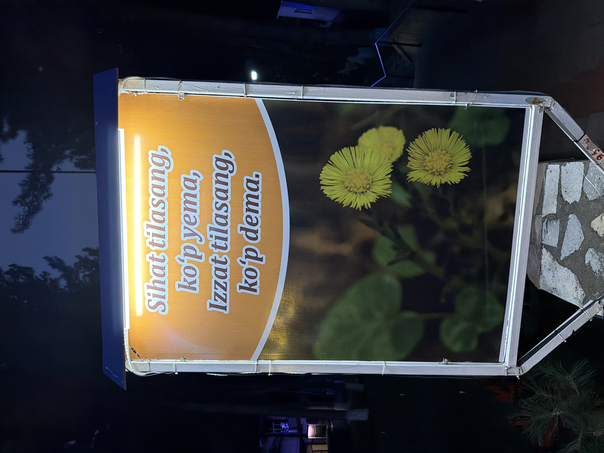 The image shows a roadside advertisement in Khumsan, Uzbekistan, at night. The advertisement features a bright orange background with white text and a photograph of yellow flowers. The text, in Uzbek, reads "Sihat tilasang, ko'p yem", meaning "If you want health, don't eat much", and "Izzat tilasang, ko'p dema", meaning "If you want respect, don't talk much". The advertisement is illuminated by a strip of LED lights above it, and it is supported by a metal frame. In the background, there are trees and buildings, suggesting an outdoor urban setting. The overall lighting is dim, typical of nighttime.