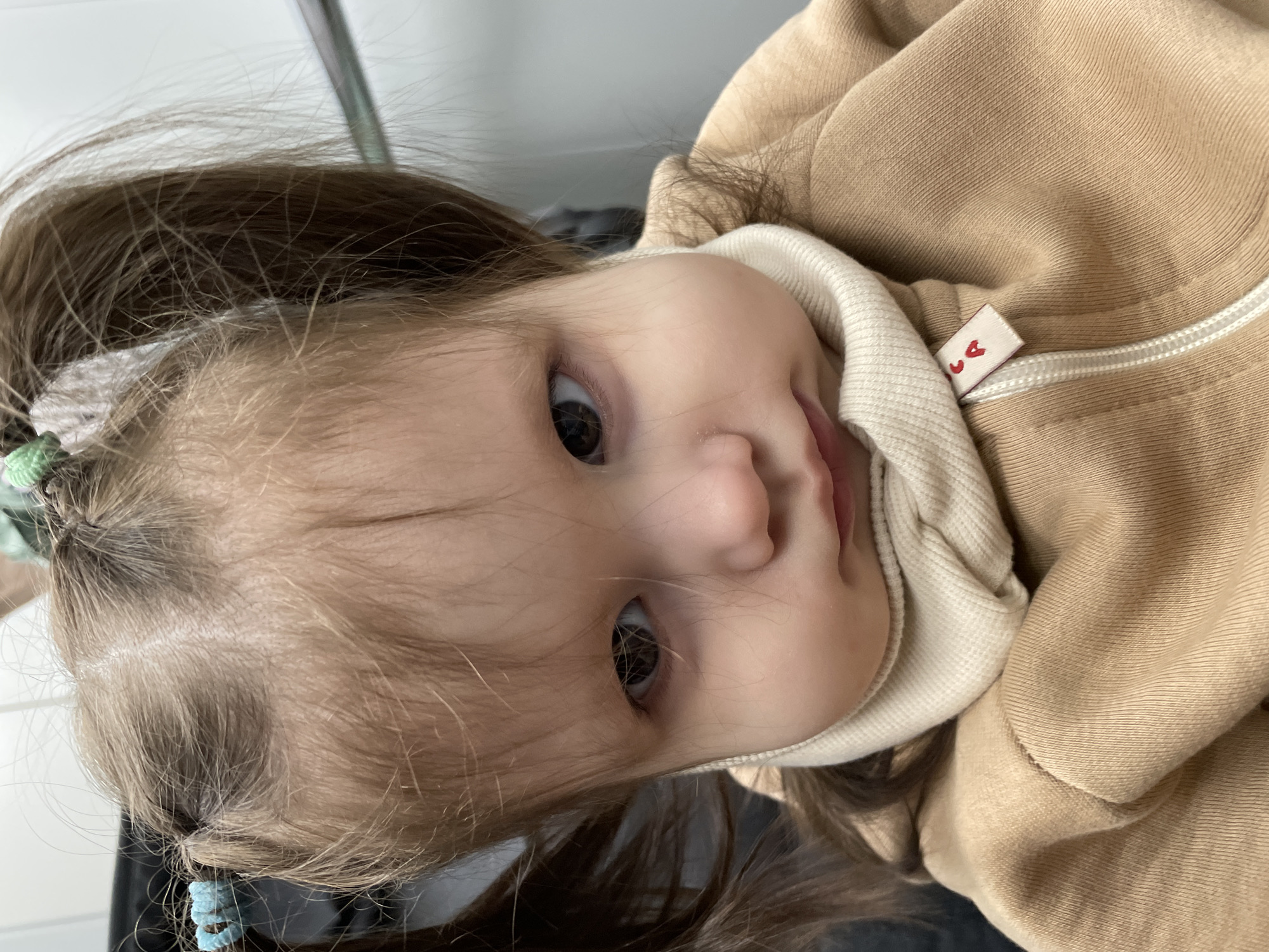 A young child with light brown hair, styled in pigtails held with light blue hair ties, is shown wearing a tan-colored hooded sweatshirt with a white-colored inner collar. The child is looking at the camera with dark eyes. A tag with a red heart symbol is visible on the sweatshirt. The environment appears to be indoors, but details are limited. There is no indication of the location being Izhevsk, Russia.
