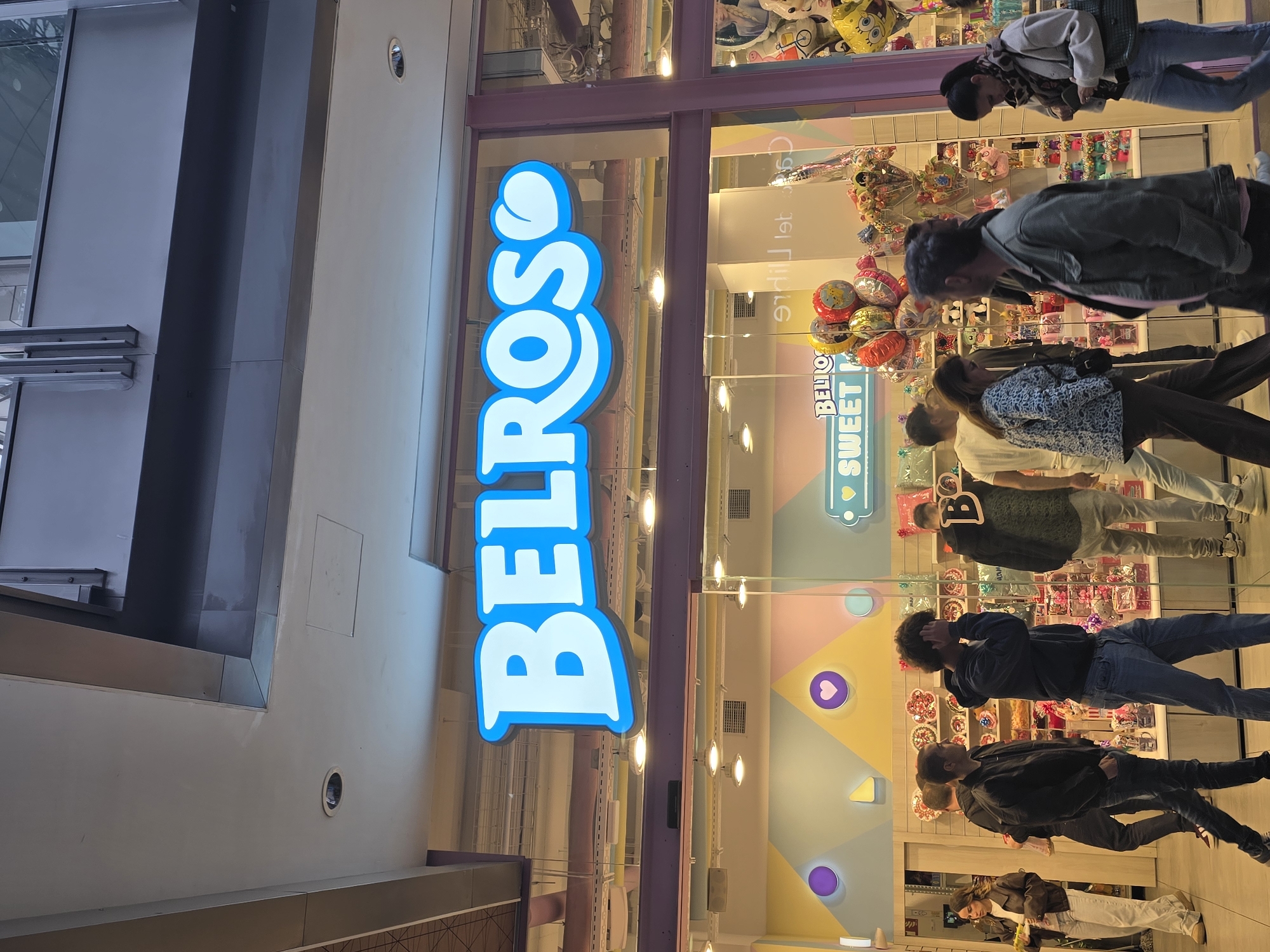 The image shows the storefront of a "Belros" store, as indicated by the large, vertically oriented, blue and white sign. The store appears to be located in a mall or indoor shopping center, evidenced by the interior view and the surrounding architectural elements. The interior of the store is filled with balloons, candy, and various other items. Several people are visible walking past the store window. The location may be in el Prat de Llobregat, Spain, though this is impossible to verify.