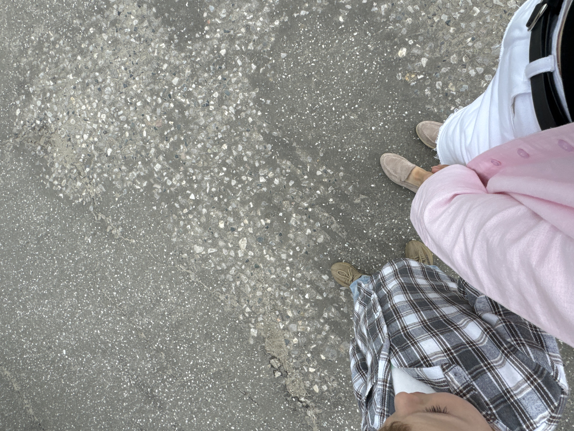 The image shows a top-down view of two individuals walking on a grey, asphalt surface with visible aggregate. The person on the left wears a plaid shirt and jeans, with their head and shoulder visible. The person on the right wears light pink linen pants and white pants with a black belt. Their feet are visible, both wearing light-colored loafers. The scene appears to be an outdoor urban environment, possibly a street or sidewalk. The location is not definitively Sharya, Russia, as there are no specific landmarks or indicators in the image to confirm this.