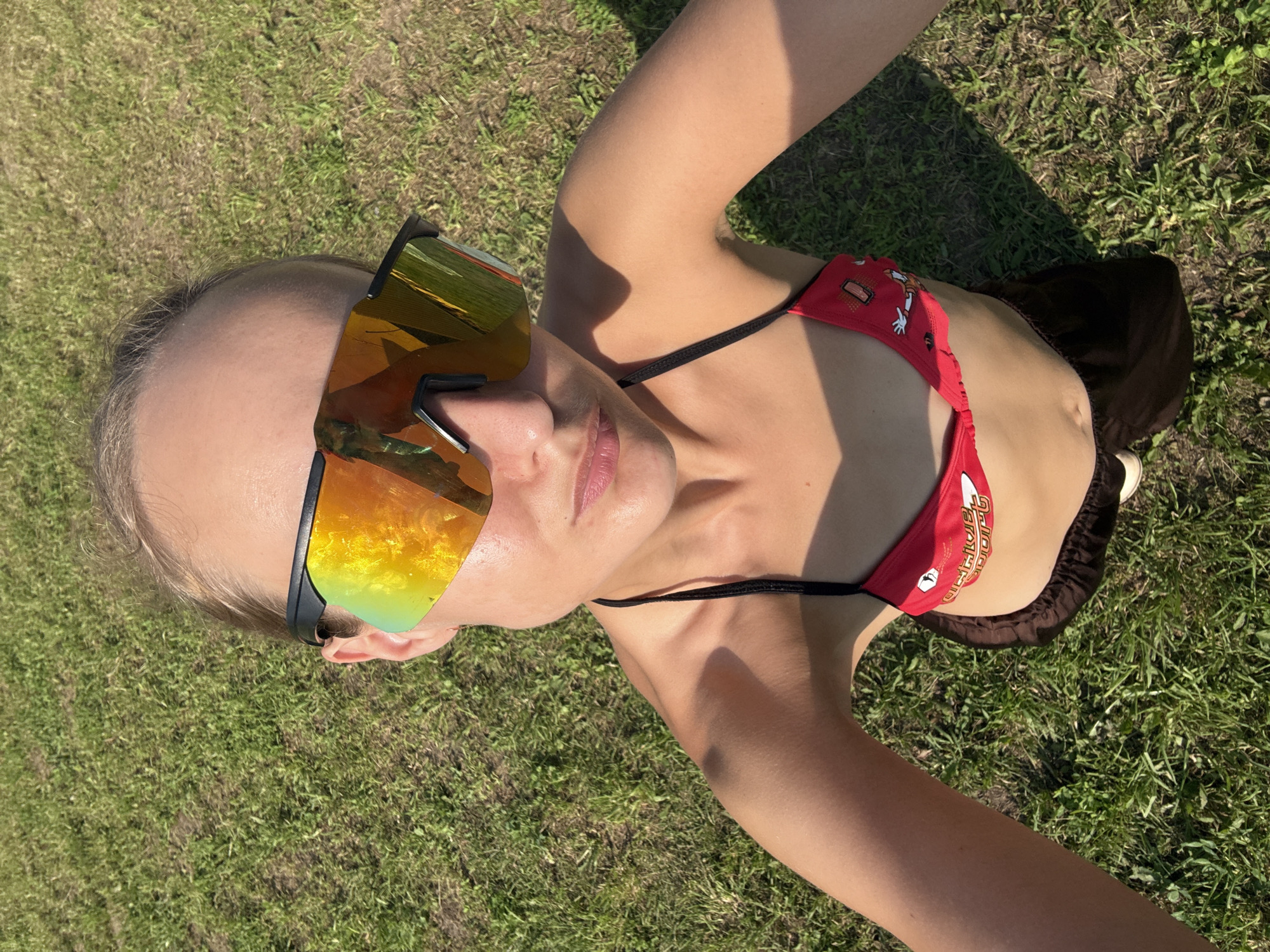 The image shows a woman lying on a grassy field, likely in Подшивалово, Russia. She is wearing large, mirrored sunglasses with a golden-orange tint and a red bikini top with a cartoonish print. Her body is angled, and her head is turned towards the camera. The sun is casting a shadow on the grass, suggesting it is daytime. The overall scene depicts a relaxed, outdoor setting, possibly a picnic or sunbathing. The image is a close-up shot, focusing on the woman and her attire against the backdrop of the green grass. There is text visible on the bikini top, which appears to be a brand name or design element, but it is largely obscured and difficult to read clearly. The lighting suggests it is a sunny day.