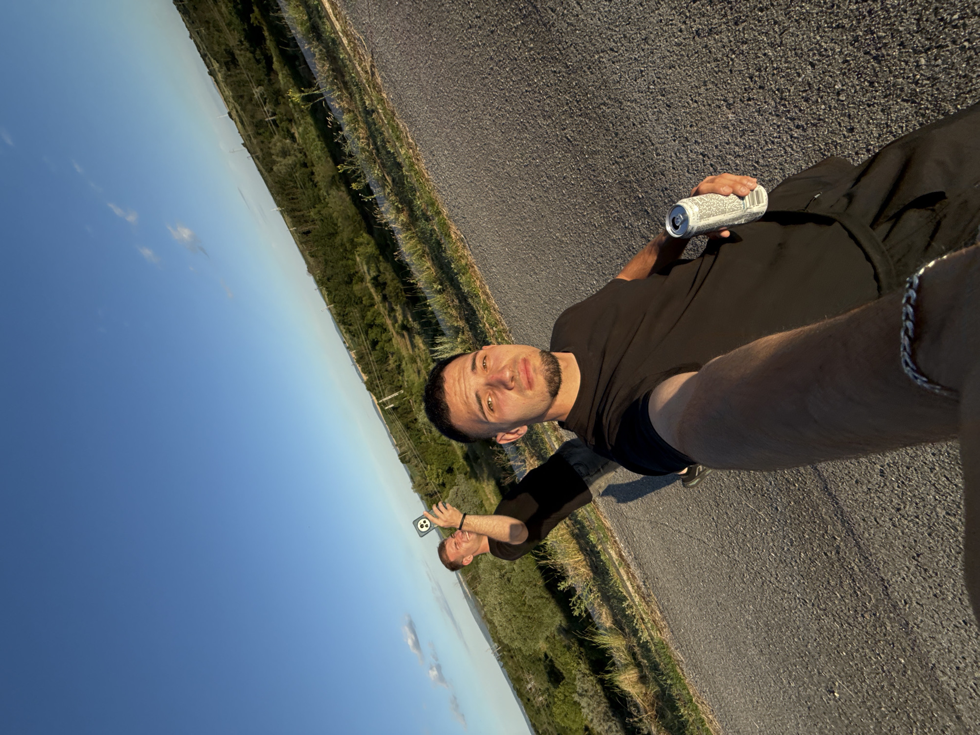 The image shows two young men on a paved road with a natural landscape of green vegetation under a clear blue sky. The man in the foreground is holding a camera or phone, taking a selfie. He is wearing a black t-shirt and dark shorts and has a beard. In his right hand, he holds a can. The second man, in the background, is also wearing a black t-shirt and is holding a phone up to take a picture. The scene appears to be an outdoor setting, possibly a park or a scenic overlook, with rolling hills and some power lines visible in the distance. The lighting suggests it is either early morning or late afternoon. The specific location in Shumen, Bulgaria, is not identifiable from the visual elements alone.