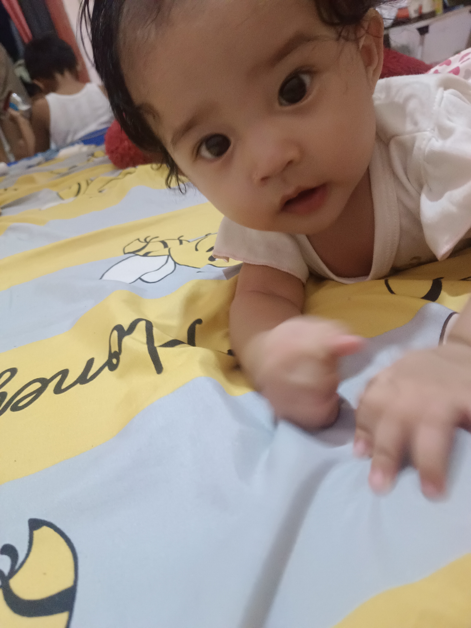 The image captures a close-up, low-angle shot of a baby lying on their stomach on a bed with a Winnie the Pooh themed blanket. The baby's face is partially visible, with dark hair and large, dark eyes looking towards the camera. Their hands are extended forward, appearing to be reaching for something. The blanket features yellow and light blue stripes, with illustrations of Winnie the Pooh and the word "honey" written in a black script. In the background, another child in a white tank top is visible, also lying down and looking away from the camera. The environment appears to be indoors, likely a bedroom, given the bed and blanket. The lighting is soft, creating a warm and intimate atmosphere. The location is identified as Kawua, Indonesia.