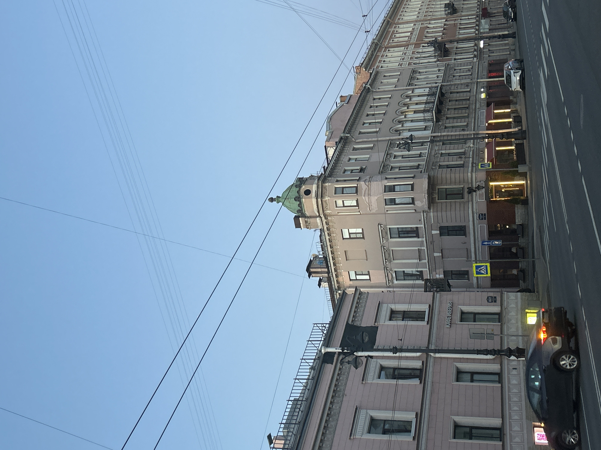 This media file captures a low-angle view of a multi-story building with a street and a dark-colored car in the foreground. The building appears to be old, with classic architectural details such as arched windows and ornate cornices. A distinctive green dome tops one section of the building, adding a touch of historical grandeur. The building's facade is a pale pinkish-gray, punctuated by numerous windows, some of which are illuminated with warm yellow light, suggesting the presence of shops or businesses on the ground floor.
Several streetlights line the sidewalk in front of the building. The street itself is paved and marked with white lines, indicating lanes for traffic. The dark car, a modern sedan, is parked on the right side of the street, its hazard lights flashing. The scene is set against a clear, pale blue sky, indicating it is daytime, likely late afternoon or early morning given the gentle lighting. Power lines crisscross the sky, a common urban sight.
There is some visible text on the building. One sign appears to say "КАФЕИВЕРИЯ" (KAFEIVERIYA), which translates to "Cafe/Catering Service." Another sign above it is too small to read. A yellow pedestrian crossing sign is also visible on the sidewalk. The overall impression is of a European city street scene, possibly in a historic district, during a quiet period. No people are clearly visible in the image.