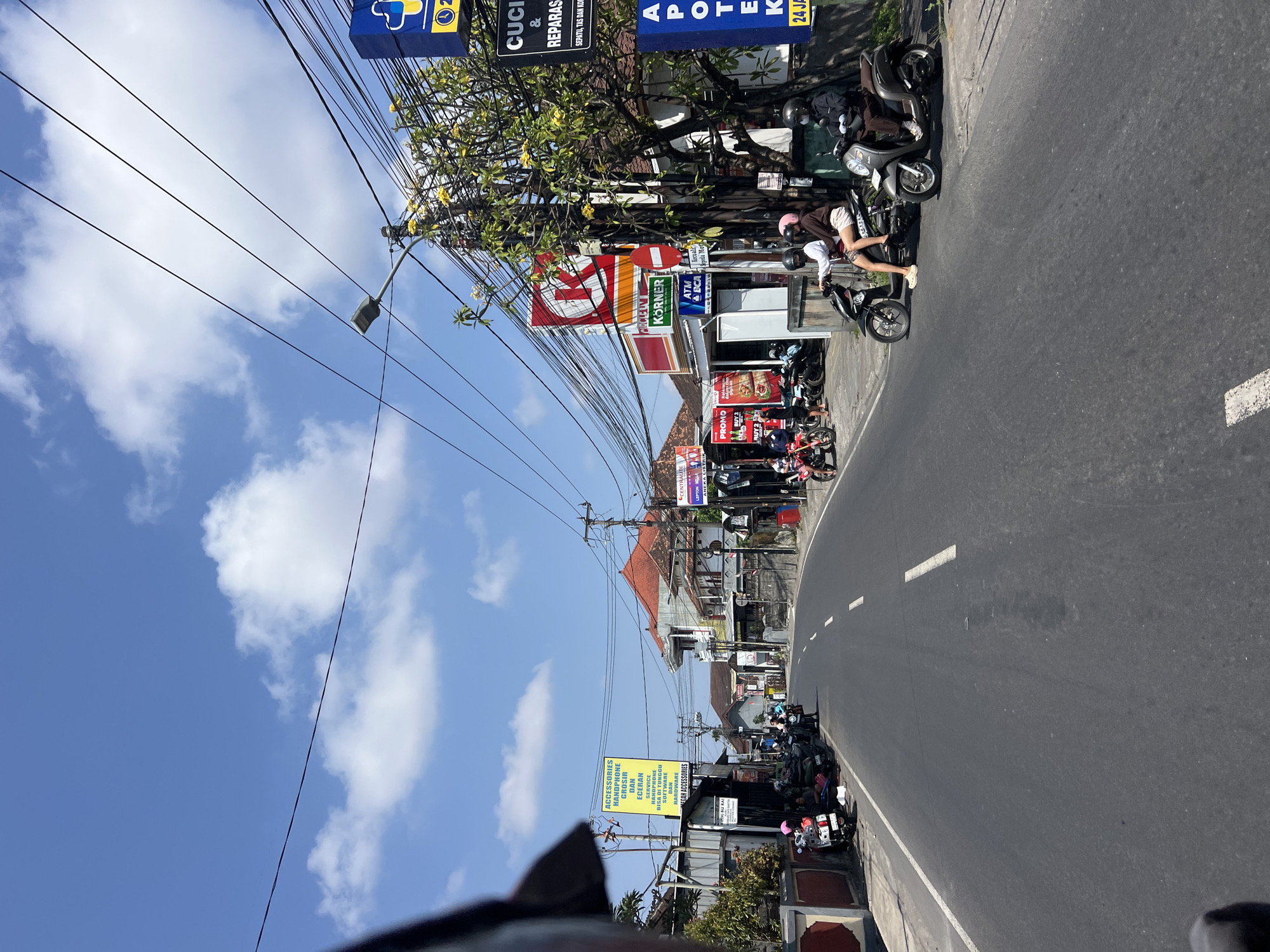 This is a ground-level, eye-level outdoor shot of a street in Denpasar, Indonesia, under a partly cloudy blue sky. The paved road has a white dashed line marking the center lane. On the left side of the road, there is a row of shops with parked scooters and motorcycles. A prominent yellow sign reads "ACCESSORIES HANDPHONE CASH DAN ECERAN SERVICE HANDPHONE BISA DI TUNGGU SEMUA UKURAN DAN MEWAH ACCESSORIES". Further down the street, more shops are visible, including a Circle K convenience store with a red and white logo and a sign that says "KORNER". There's also a blue sign with a yellow plus symbol and a clock face, indicating a pharmacy or clinic. Several people are on or near motorcycles. On the right side of the road, there's a tree with yellow flowers, a utility pole with numerous wires, and signs for "CUCI & REPARASI SEPATU, TAS DAN DOMPET" and "APOTEK K". A few people are riding scooters and motorcycles. The overall atmosphere is that of a bustling street in a tropical urban environment.