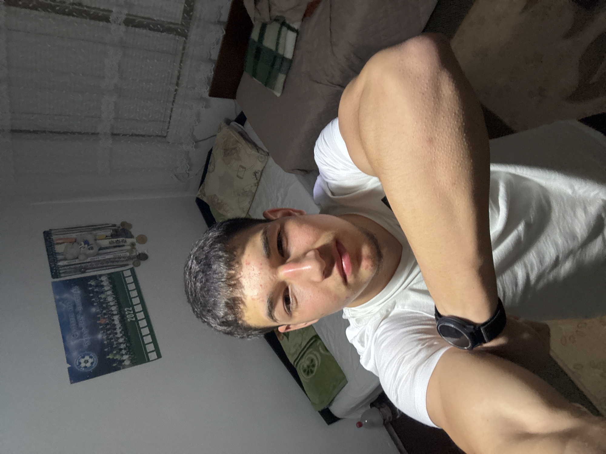 A selfie from the chest up shows a person with short dark hair, light skin, and a white T-shirt with a black smartwatch on their left wrist. The background shows a bedroom with a bed and a window with lace curtains. There is a poster on the wall. The location is possibly a private residence in Zlatograd, Bulgaria.