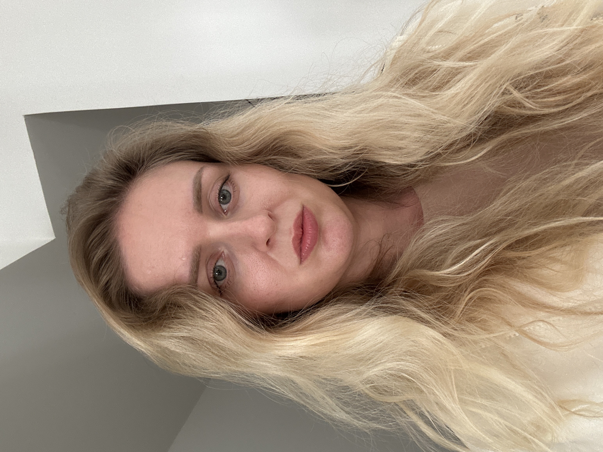 A fair-skinned woman with long, blonde wavy hair is positioned close to the camera, tilting her head slightly to the right. She has light eyes and pink lips. The background consists of white and gray surfaces suggesting an indoor setting, possibly a room with walls and ceiling. No specific location in Petah Tikva, Israel is discernible. There are no visible actions or interactions.