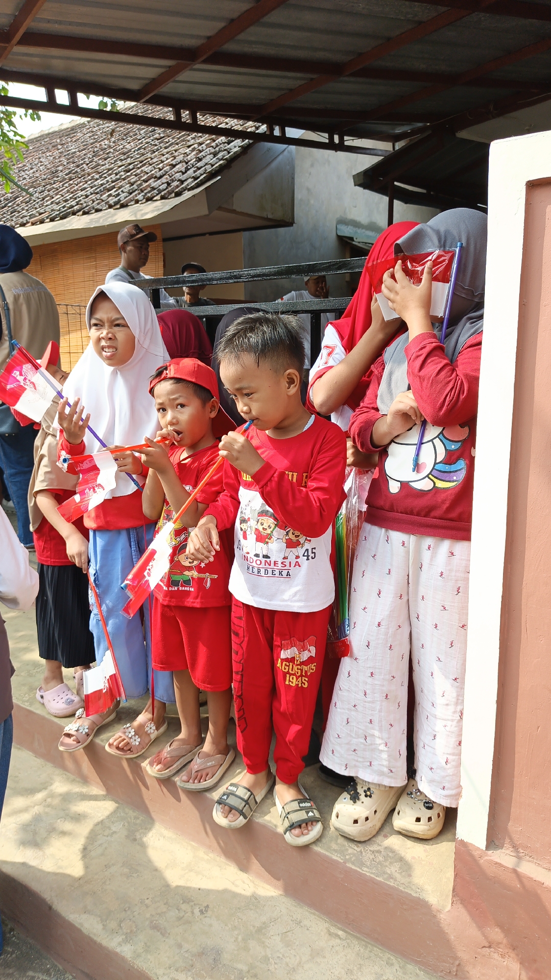 In Linggasirna, Indonesia, a group of children are standing on a concrete step, holding small Indonesian flags. The children are dressed in red and white clothing, with some wearing red shirts and pants, and others wearing white shirts with red accents. The flags are red and white, representing the Indonesian flag. The background shows a building with a tiled roof and a shaded awning. The scene appears to be outdoors on a sunny day, possibly during a national celebration or event. The children are looking in different directions, some engaged in conversation or looking at the flags they are holding. The overall impression is of a cheerful gathering of children participating in a patriotic event.