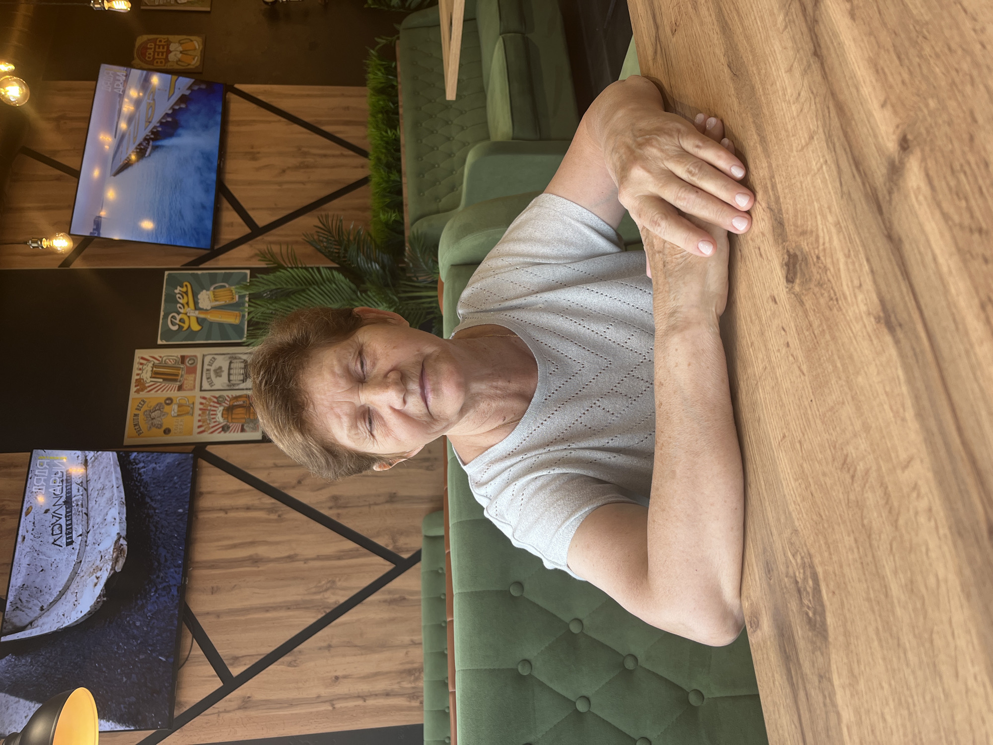 An elderly woman with short brown hair and closed eyes sits at a wooden table. She is wearing a light gray, short-sleeved shirt with a diamond pattern. Her arms are resting on the table, with her hands clasped. Behind her, a green tufted booth seating area is visible. The background features wooden paneling on the walls, with black geometric accents. Two television screens are mounted on the wall, displaying images. One screen shows a close-up of a damaged industrial object with the word "ADVANCING" on it. The other screen displays a pier extending into the water. Decorative light bulbs hang from the ceiling. The overall setting appears to be a modern cafe or restaurant.