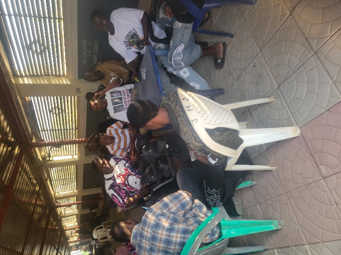 Seven adult males are seated around a rectangular blue table on a tiled floor under a corrugated metal roof structure. The scene is located in a covered outdoor or semi-outdoor area within the city of Ibo, Nigeria.
**People:**
- Two men are seated with their backs to the camera in the foreground: one on a green plastic chair wearing a blue plaid shirt and dark trousers, and another on a white plastic chair wearing a patterned top and dark trousers.
- Five men face the camera, seated around the blue table. From left to right:
- A man in a white, purple, and black patterned t-shirt.
- A man in an orange and white striped t-shirt.
- A man in a white t-shirt with a dark graphic print ("THE SPIRIT OF YOUTH...") is gesturing with his right hand.
- A man in a tan-colored hoodie.
- A man in a white t-shirt with a dark graphic (partially visible "URBAN TEMPLE") is leaning back in a blue plastic chair, smiling. He wears light-colored jeans with white patterned patches and dark flip-flops.
All individuals appear to be engaged in a social interaction or conversation.
**Objects and Environment:**
- The floor is covered with large square tiles featuring a circular, radiating pattern in shades of beige and light brown.
- The seating consists of various plastic chairs: white, green, and blue. A "J K S" marking is visible on the backrest of the white chair in the foreground.
- The overhead structure is a corrugated metal roof supported by visible beams.
- In the background, a light-colored wall with horizontal louvers or vents allows sunlight to pass through. Below this, a darker surface, possibly a blackboard or painted wall section, has faint white text, including "NO SMOKING OR... HARM DRUGS" and "ESA" on the lighter wall.
- Small, indistinct objects, possibly a green bottle or container, are visible on the blue table.
The area is illuminated by natural light, casting shadows on the floor and under the roof structure.