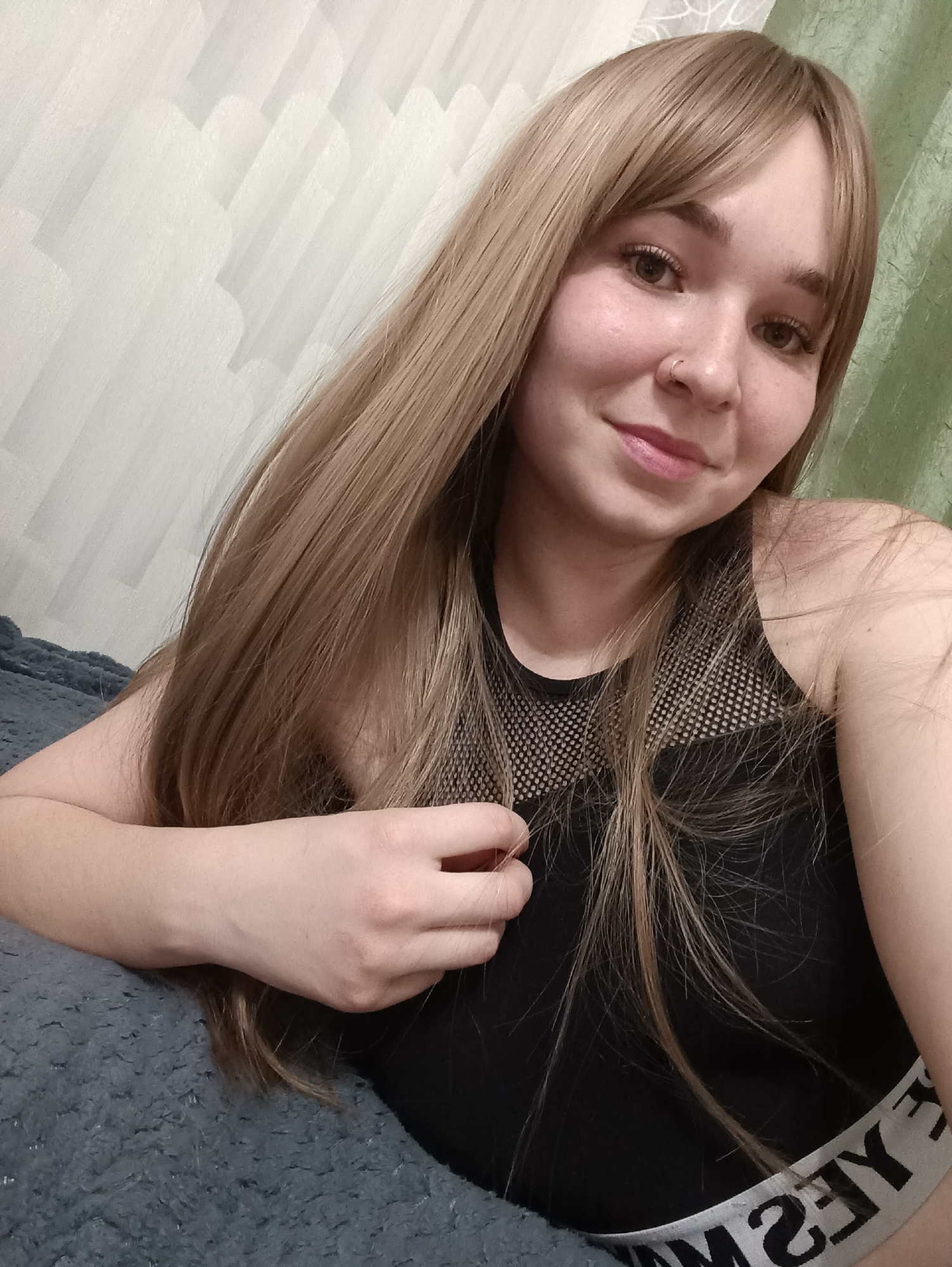 A young adult female is depicted in an indoor setting, specified as Izhevsk, Russia. No specific street or building within Izhevsk is identifiable. She has long, straight, light brown hair with bangs and displays a slight smile. A small metallic ring is present in her left nostril. She is wearing a black tank top with a mesh upper chest area and a white elastic band at the bottom featuring black, reversed text. Her left arm supports her, while her right hand is situated near her chest, touching her hair. The background behind her left side is a light-colored wall with a textured pattern of repeating rounded forms. Behind her right side, a green, vertically folded curtain is visible. She is resting on a dark blue-gray, textured fabric.